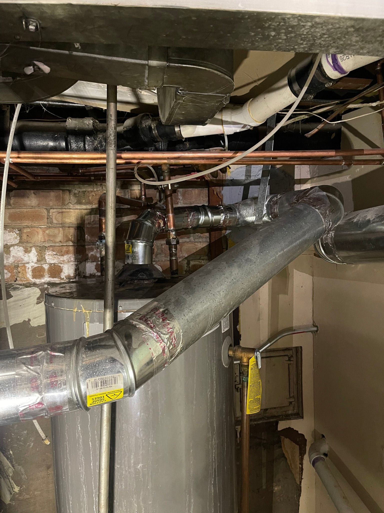 Plumbing and ductwork in a basement with a water heater, brick wall, and silver ductwork.
