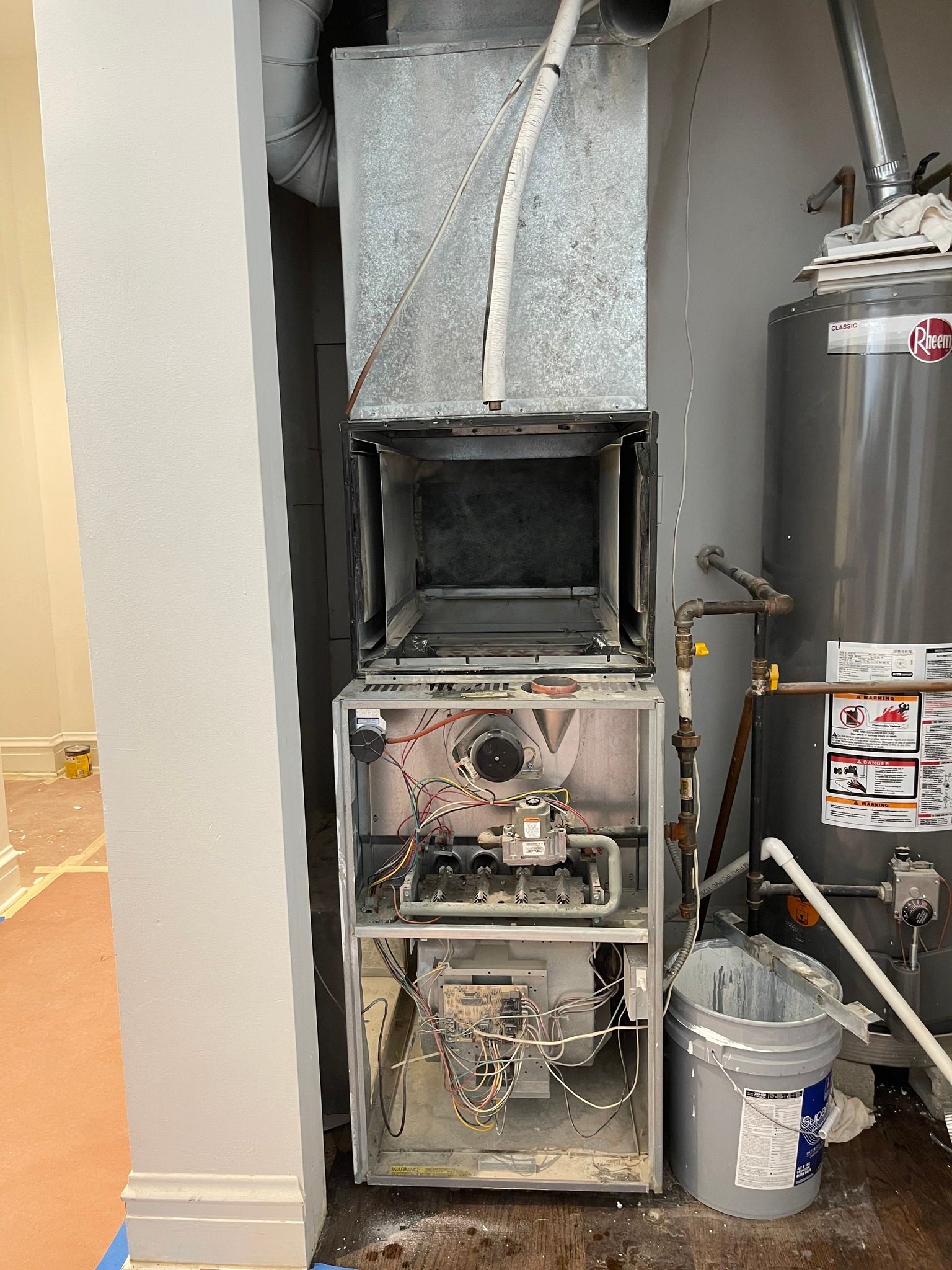 Furnace and water heater in a utility room. Furnace front is open, a bucket of paint sits nearby.
