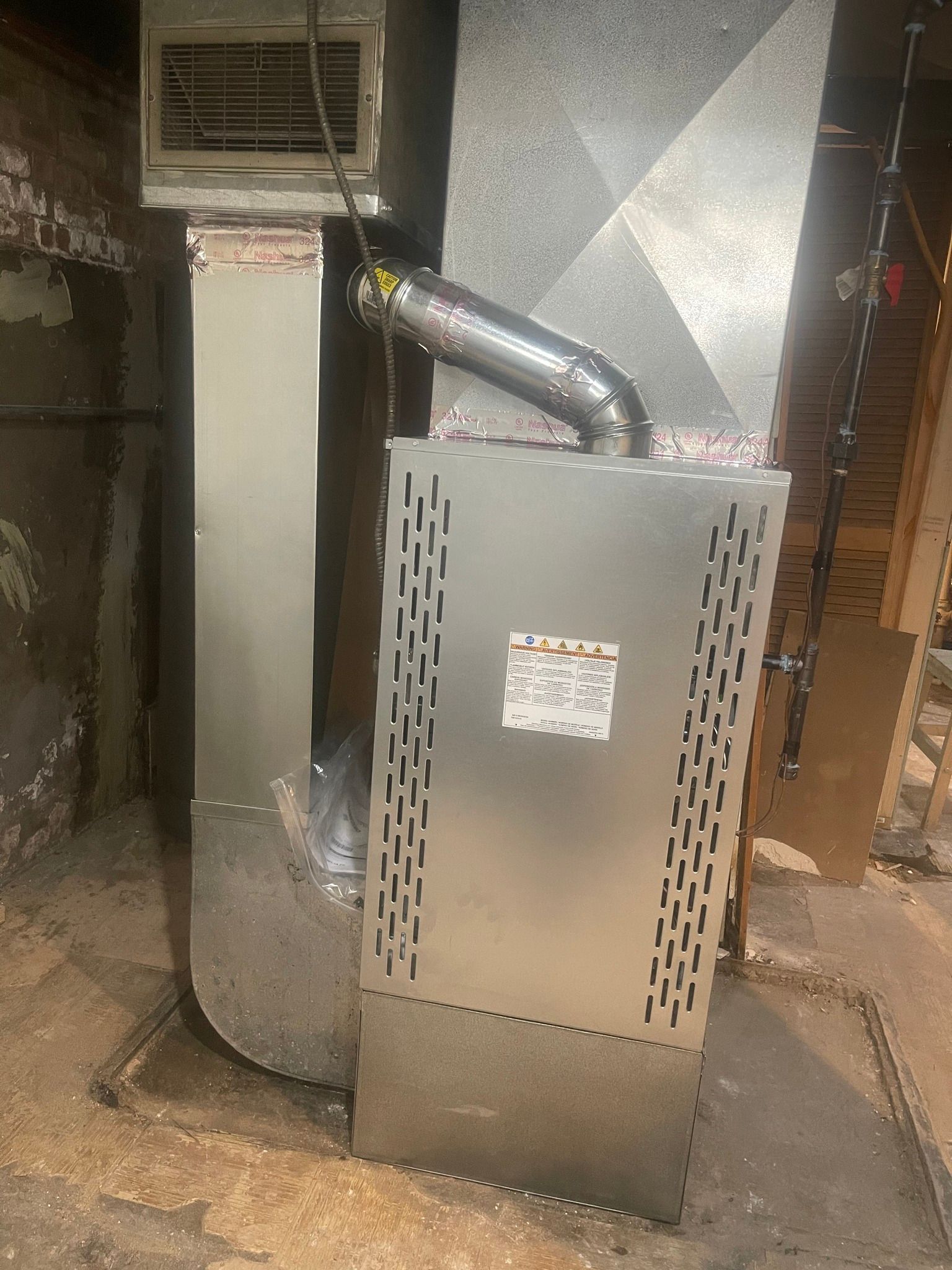 A silver furnace in a basement setting with ductwork and vent pipe attached.