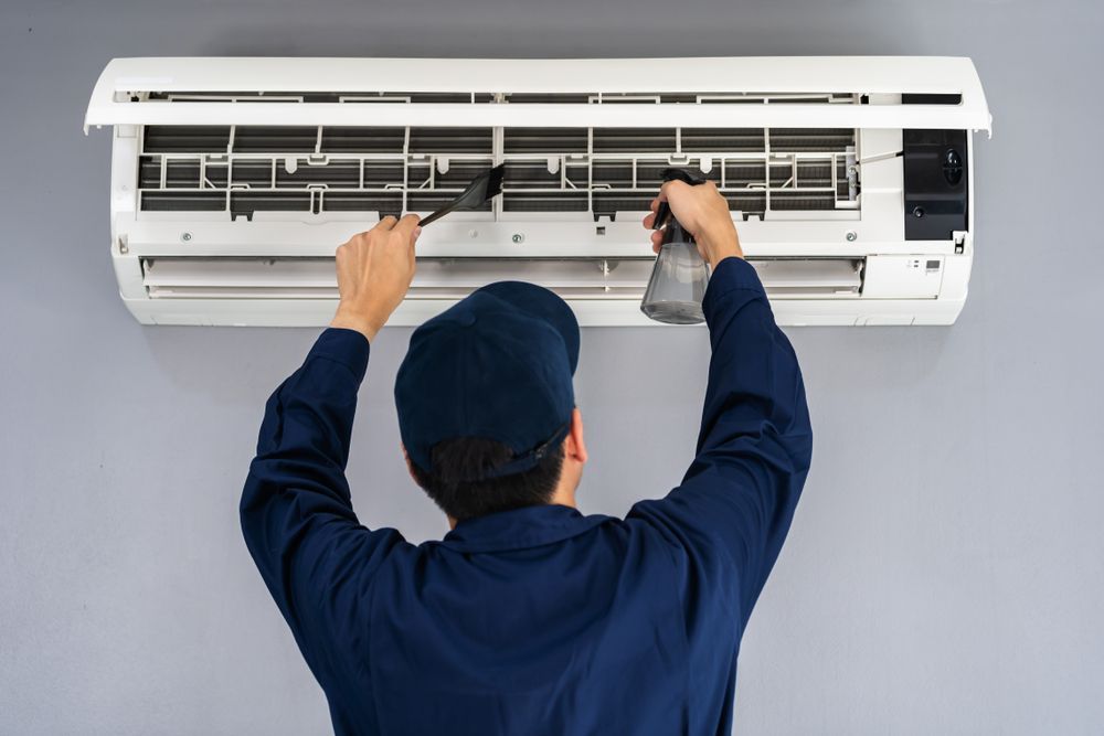 A person in a blue uniform cleans a white wall-mounted air conditioner with a brush and spray bottle.