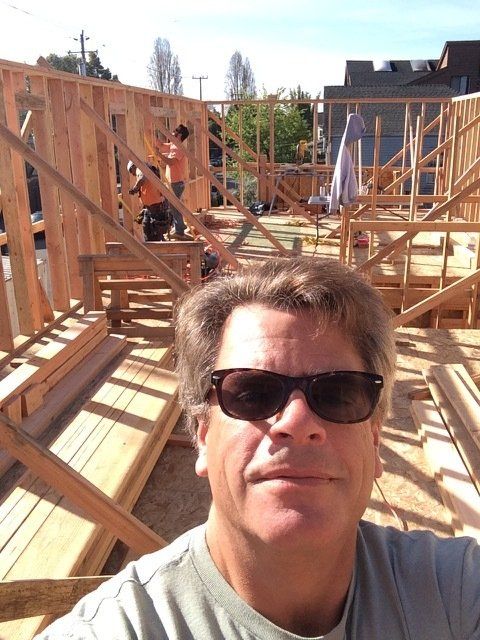 A man in sunglasses takes a selfie at a construction site with wooden wall frames and workers in the background.