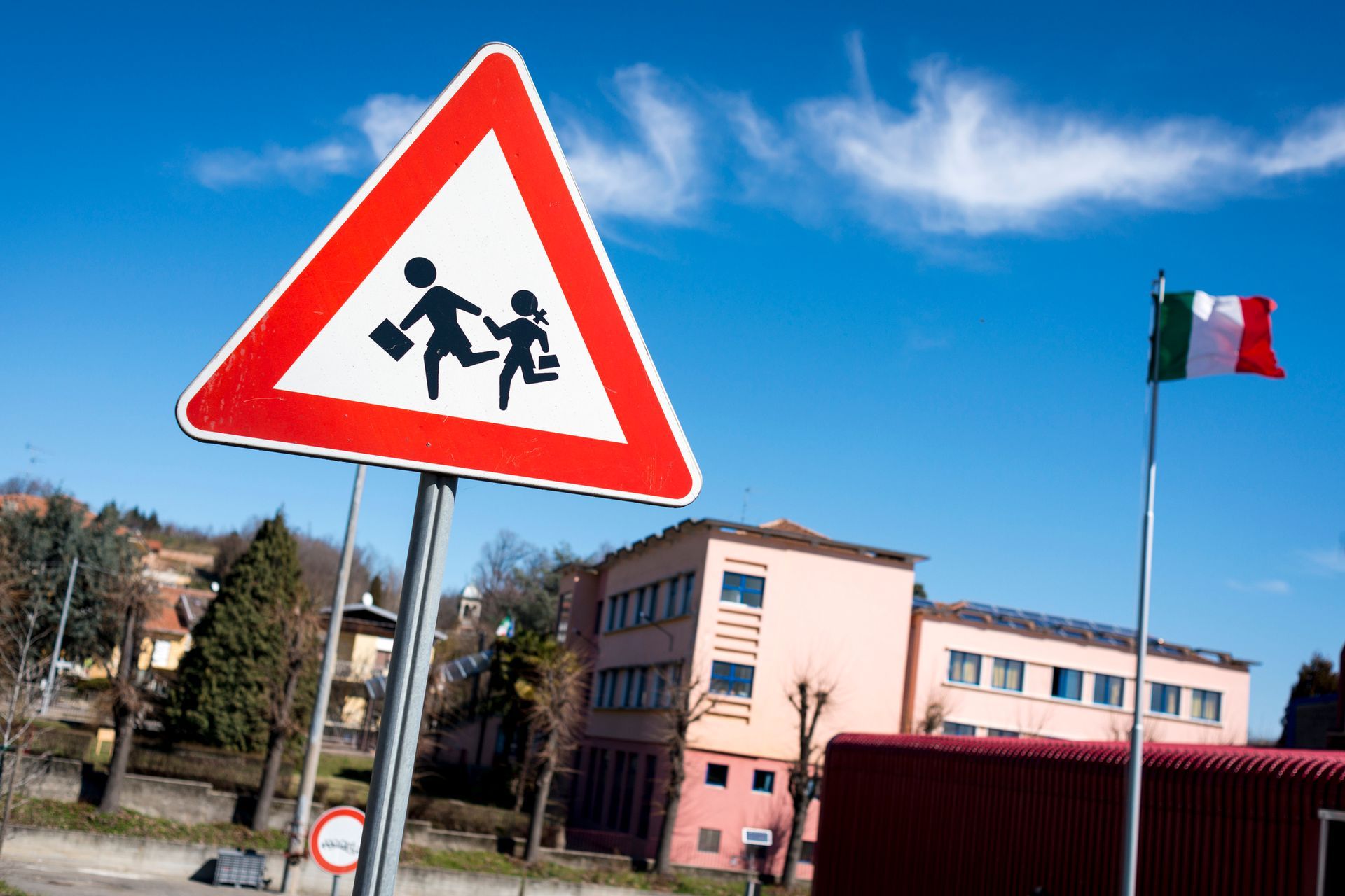 Cartello di zona scolastica con bambini, bandiera italiana, edificio e cielo azzurro.