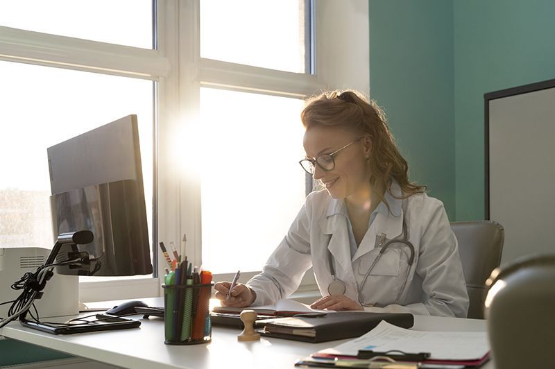 Una dottoressa, con occhiali e camice, scrive alla scrivania in un ufficio ben illuminato.