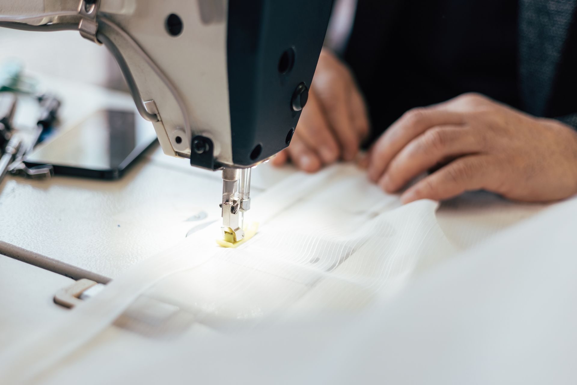 Persona che cuce con una macchina da cucire su un tessuto bianco. Sono visibili le mani.