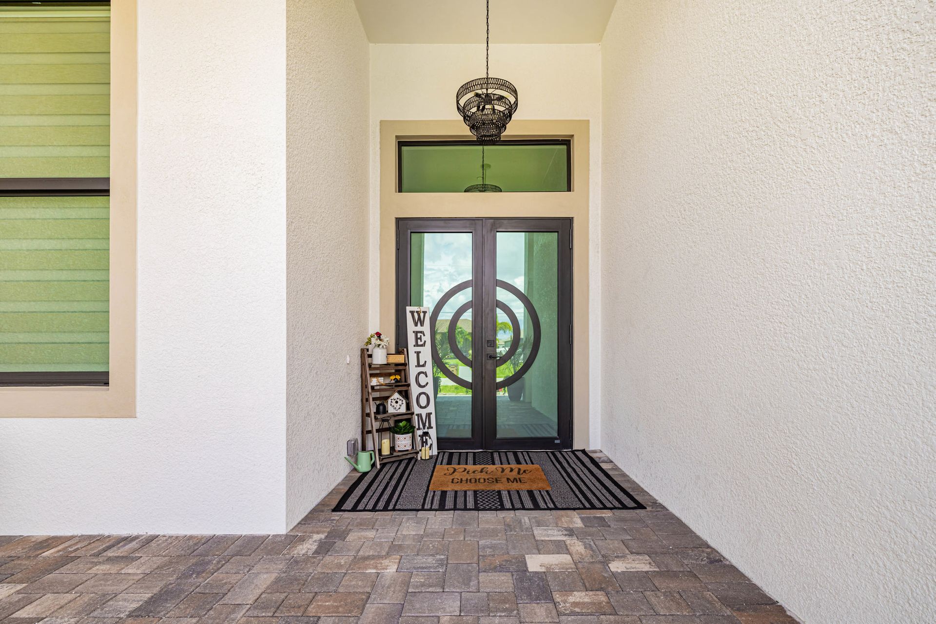 Front entrance with double doors and a welcome mat. Gray, white, and black accents. Textured white walls, decorative light fixture.