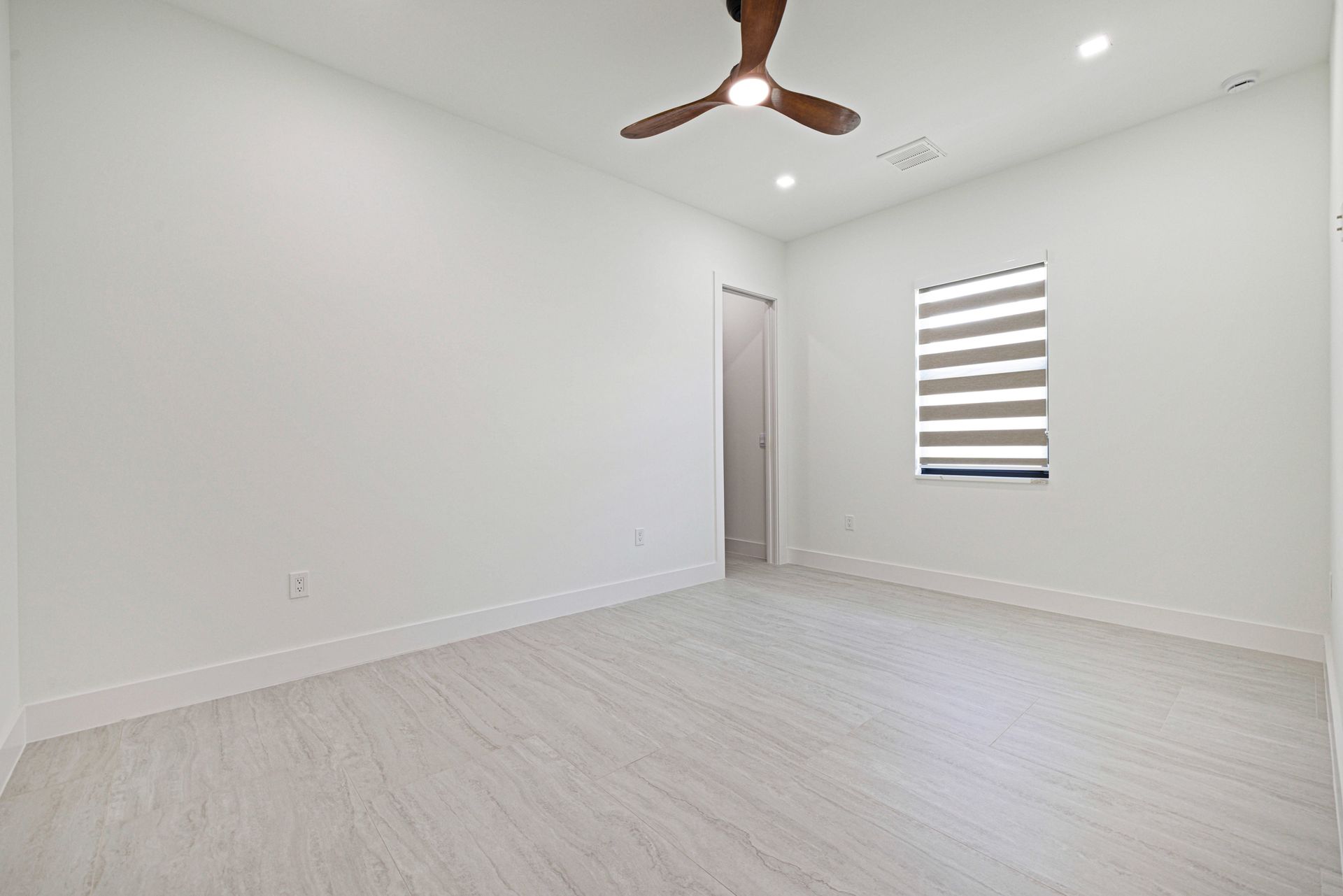Empty room with white walls, light wood-look floors, a ceiling fan, a window with blinds, and a door.