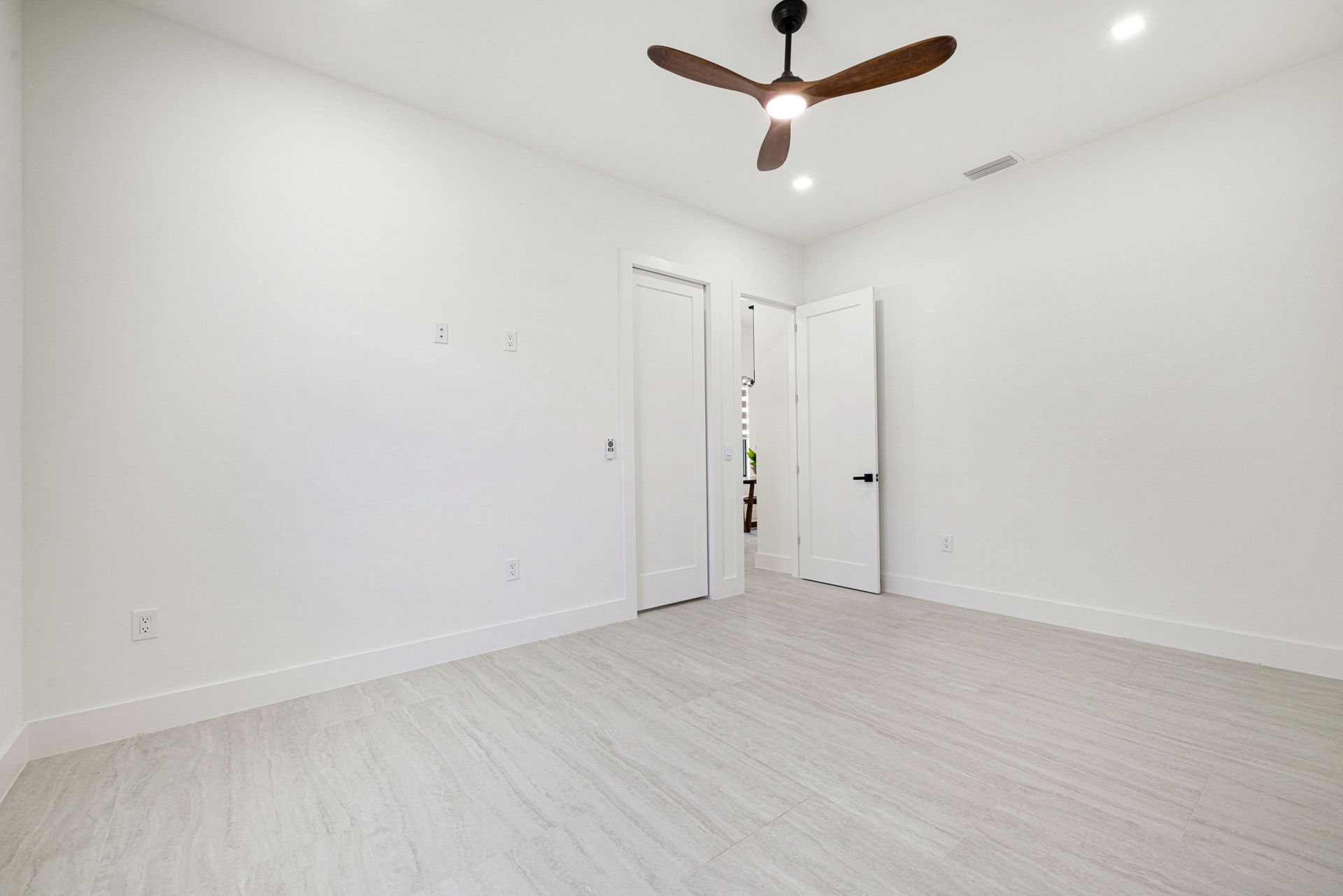 Empty room with white walls, light wood floors, ceiling fan, and two closed doors.