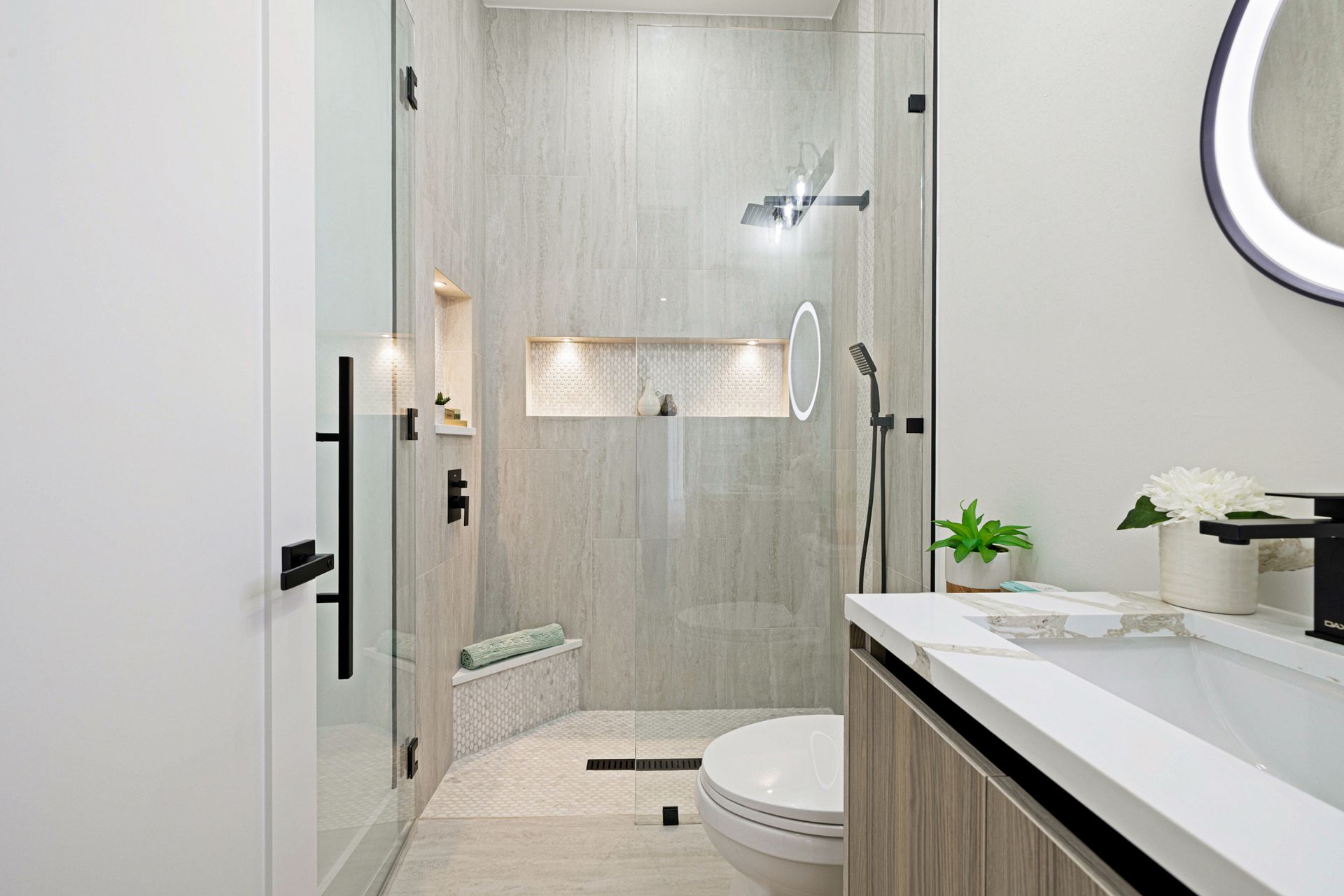 Modern bathroom with glass shower, white and gray tile, black fixtures, and a round lit mirror.