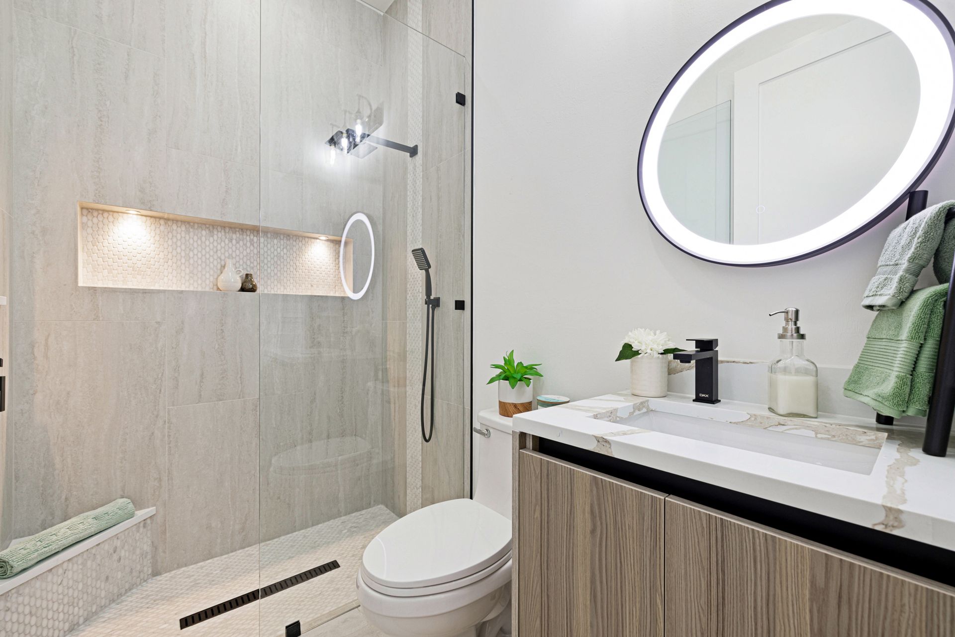 Modern bathroom with a glass shower, round illuminated mirror, and light wood vanity.