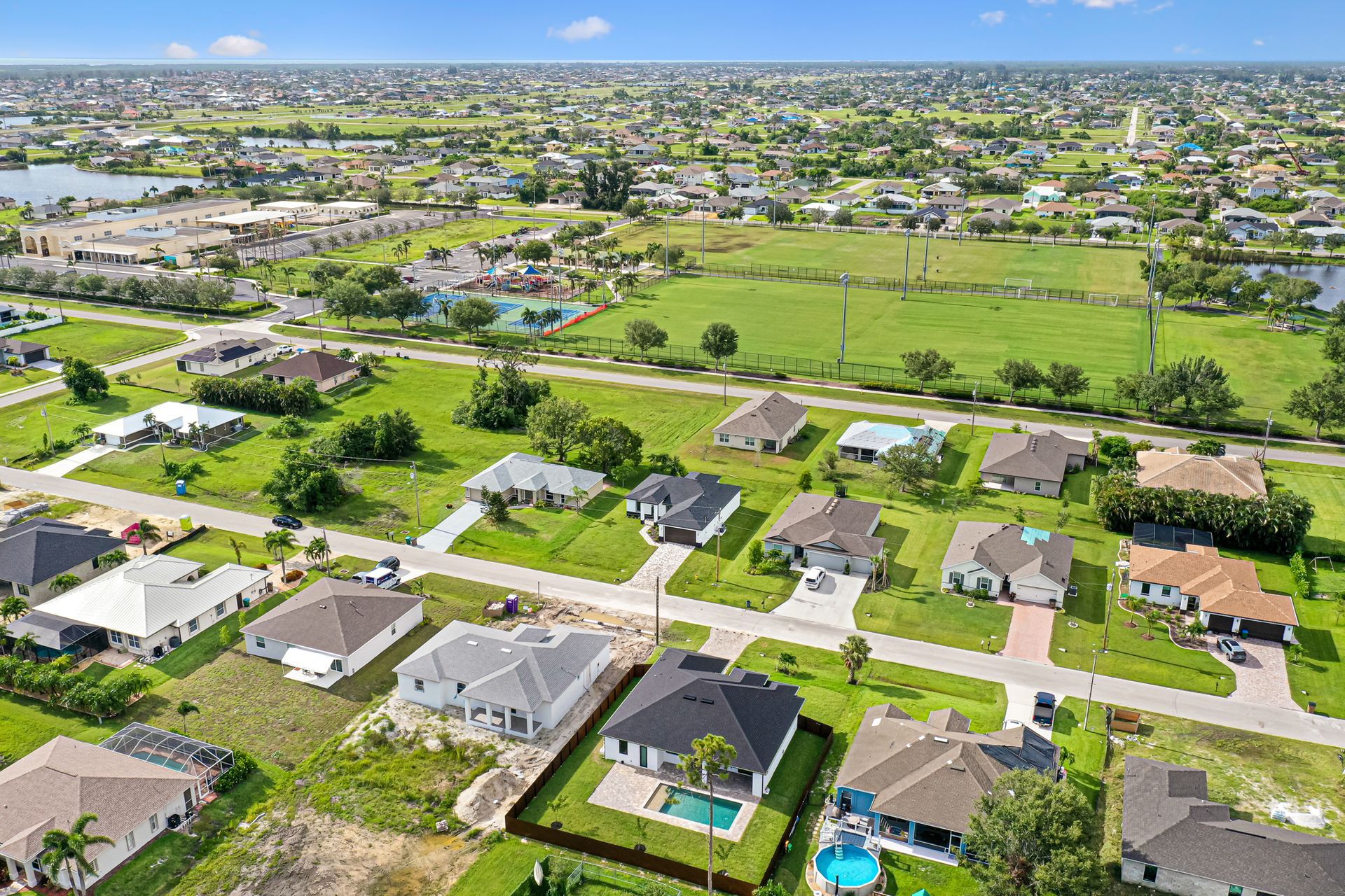 Aerial view of a suburban neighborhood with houses, roads, green spaces, and a lake under a blue sky.