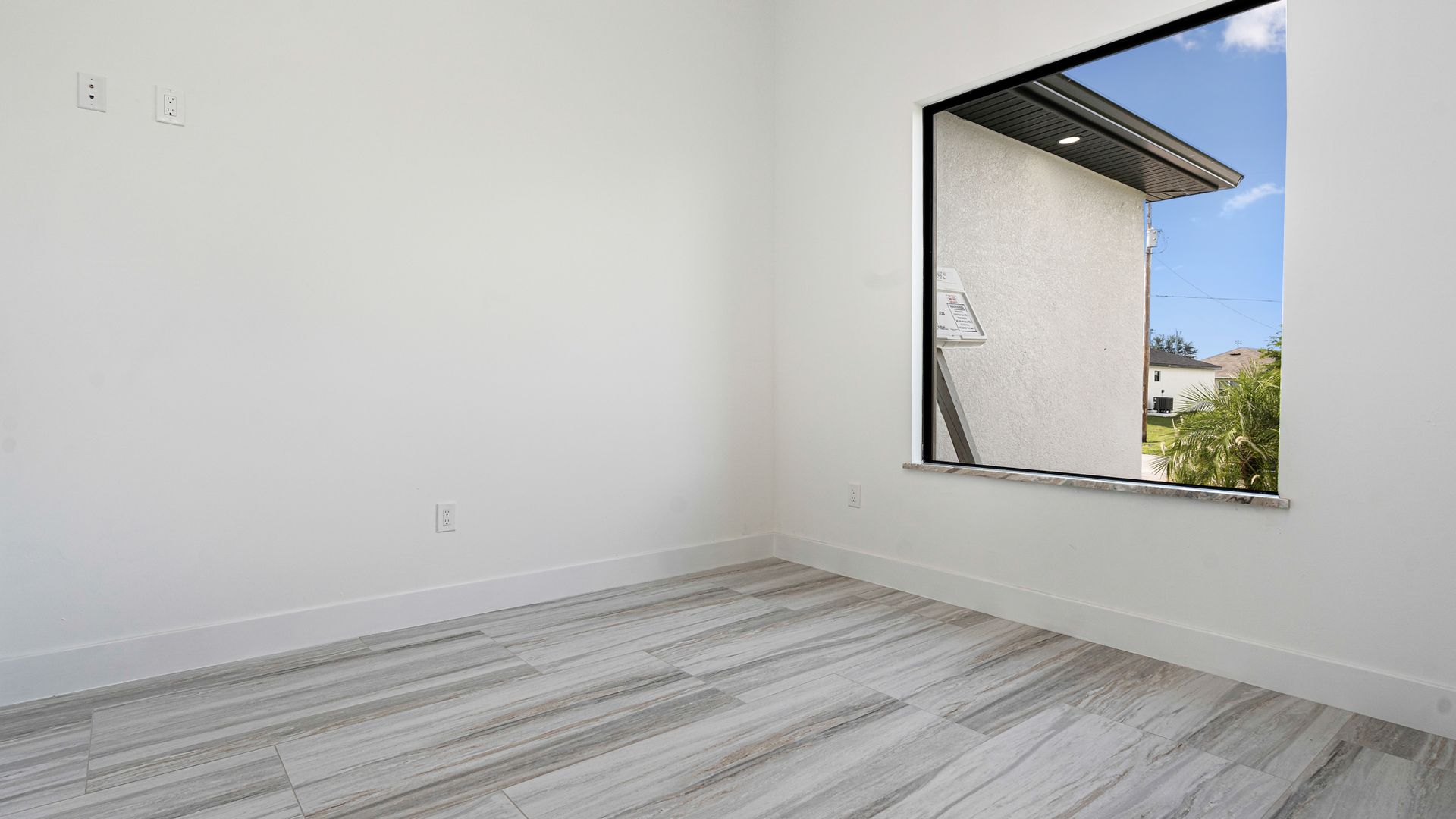 Empty room with white walls, light wood-look floor, and a window with a view of another building and blue sky.