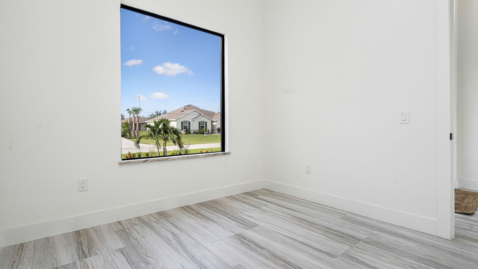 Empty room with a large window showing a blue sky and houses, white walls and gray floor.