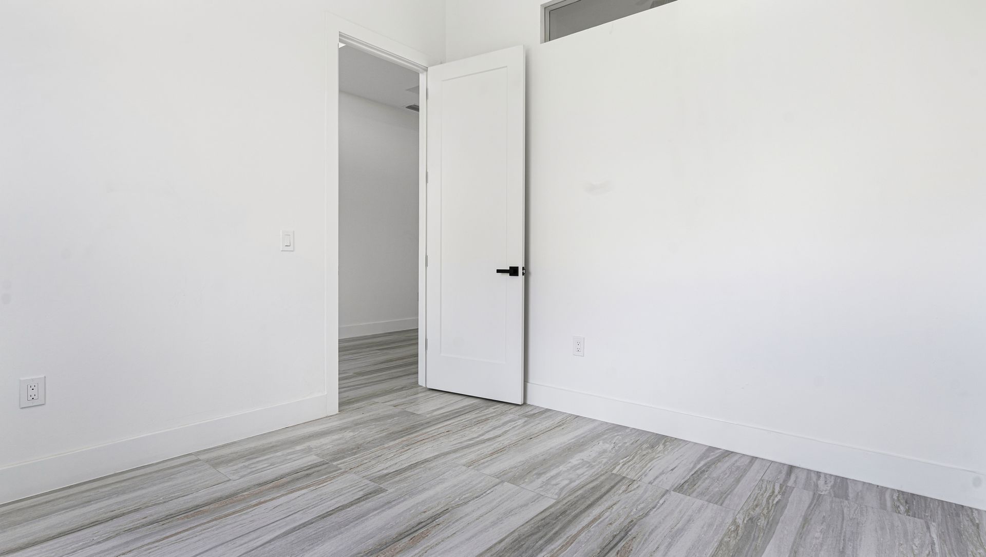 Empty room with white walls and gray wood-look flooring. A white door opens to another room.