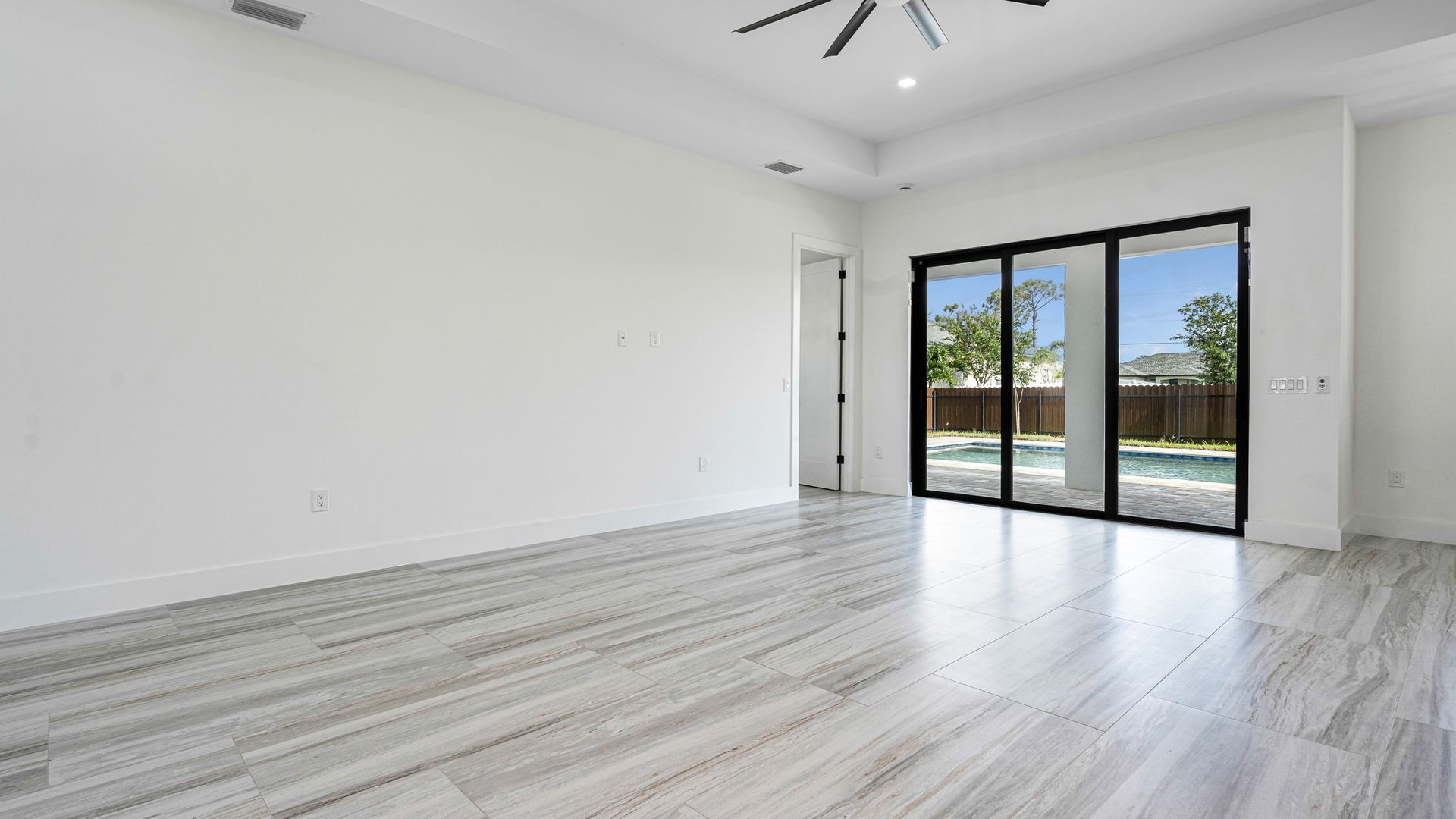 Empty room with white walls, gray tiled floor, sliding glass doors to backyard, and a ceiling fan.
