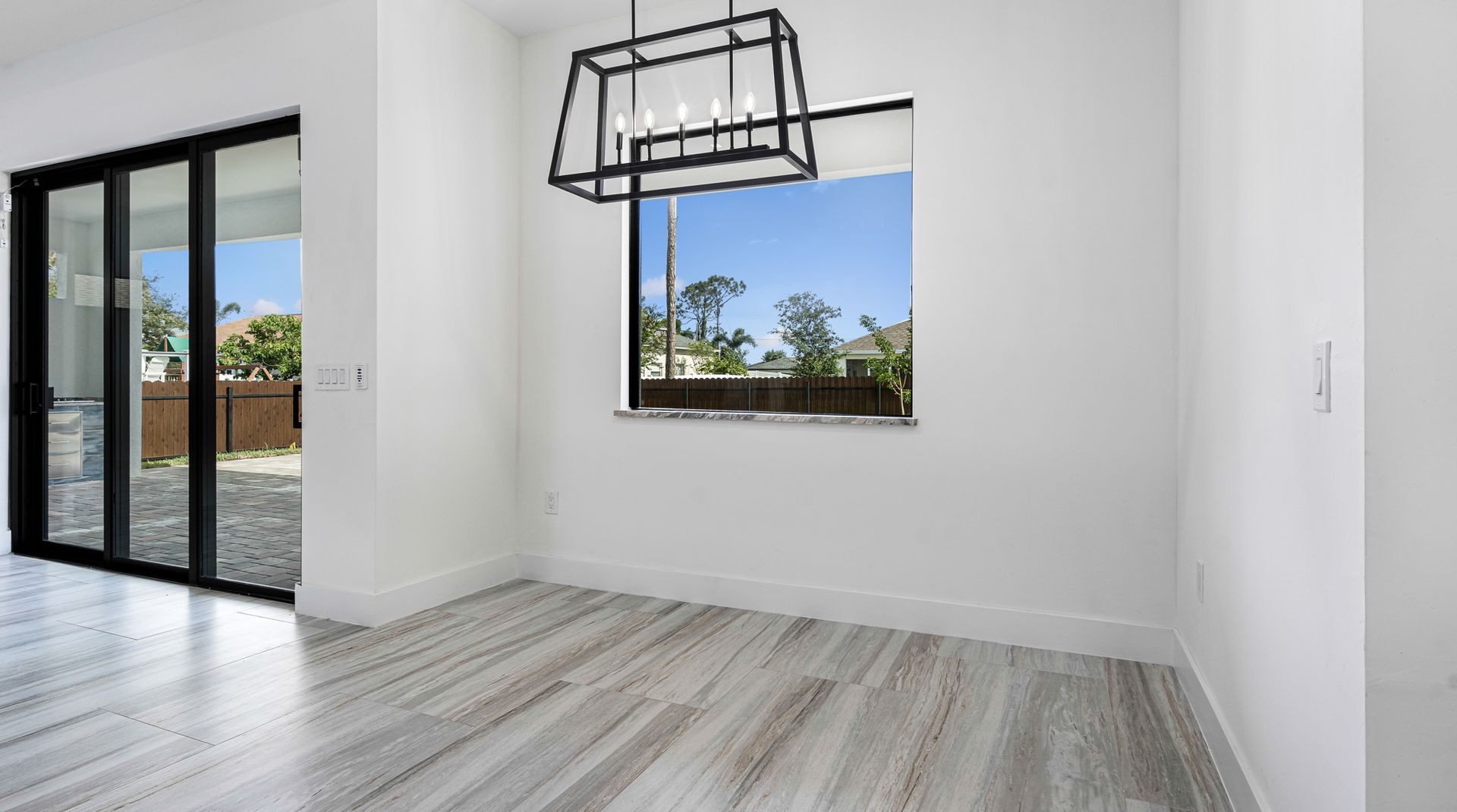 Empty dining room with light wood-look flooring, white walls, large window, and black chandelier.