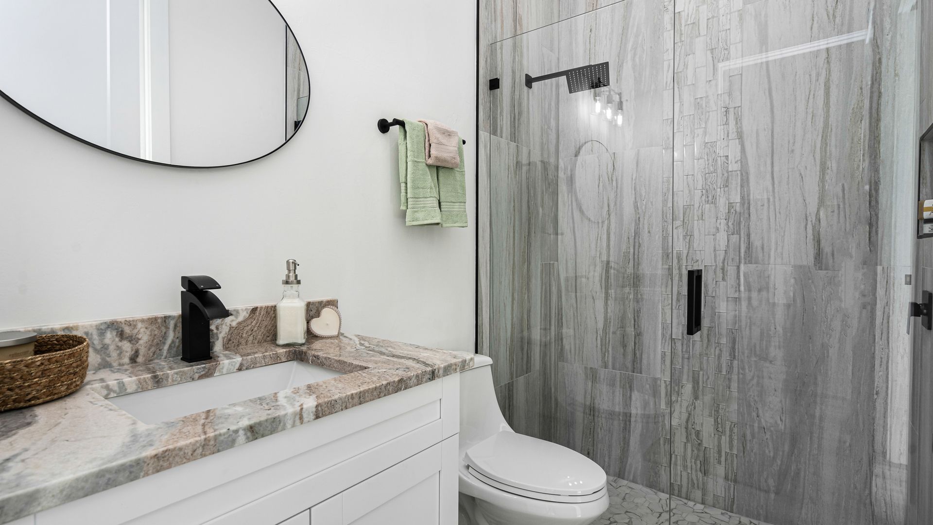 Modern bathroom with a white vanity, gray stone-look shower, round mirror, and black fixtures.