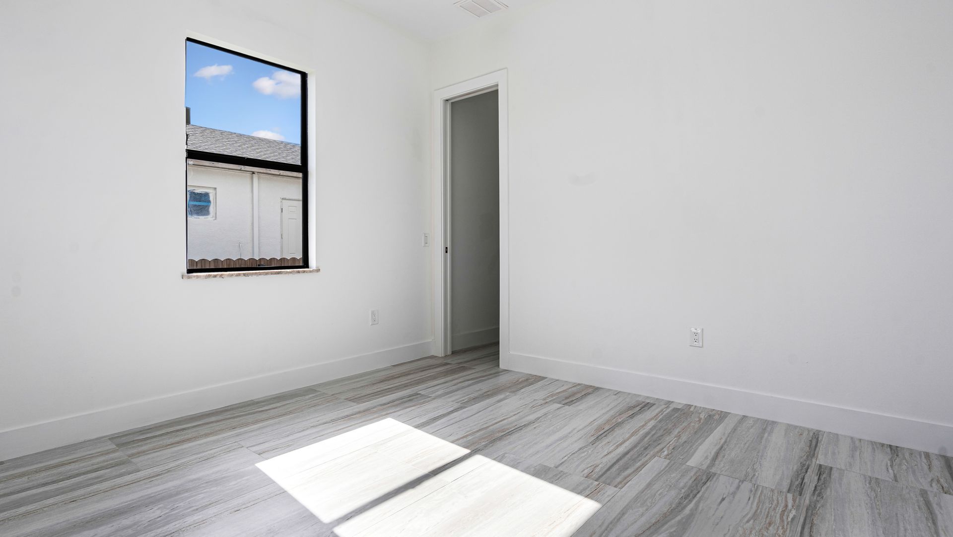 Empty, white-walled room with gray tiled floor, window showing outside buildings, and an open doorway.
