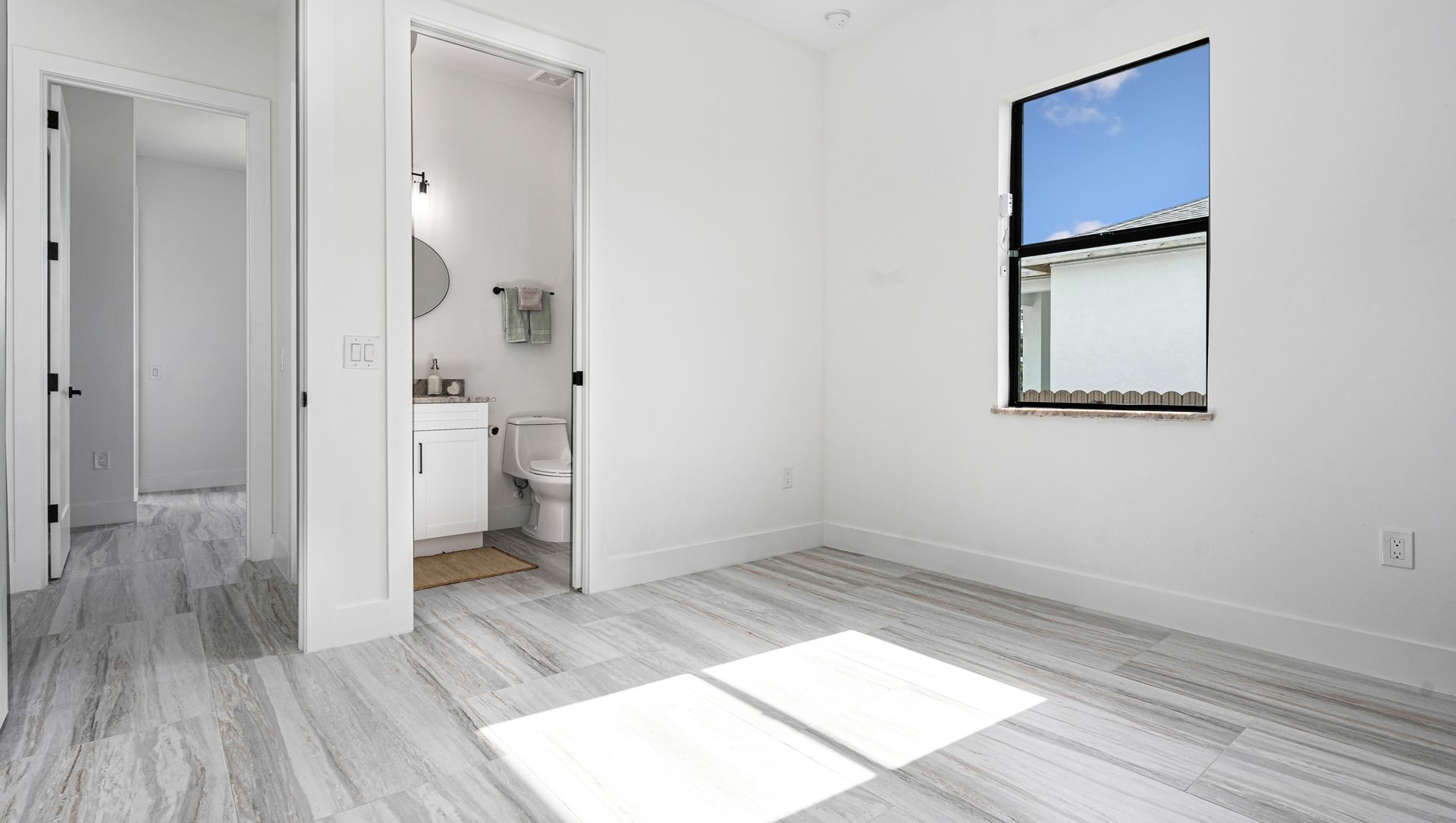 Empty room with gray wood-look floor, white walls, and a window. A doorway leads to a bathroom.