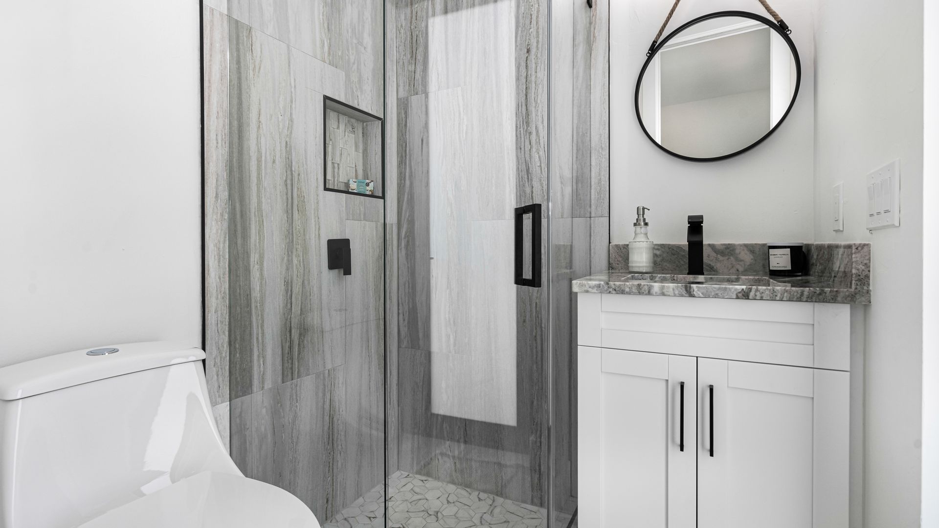 Bathroom with white toilet, glass shower, white vanity, and round mirror.