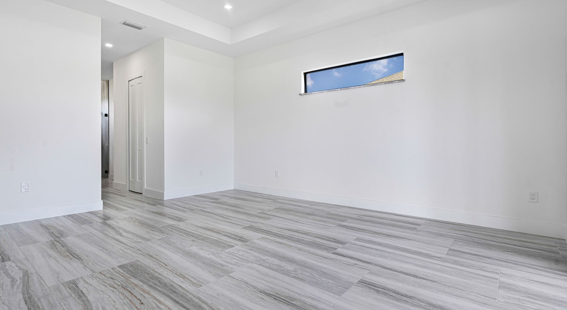 Empty room with gray wood-look flooring, white walls, and a small window showing a blue sky.