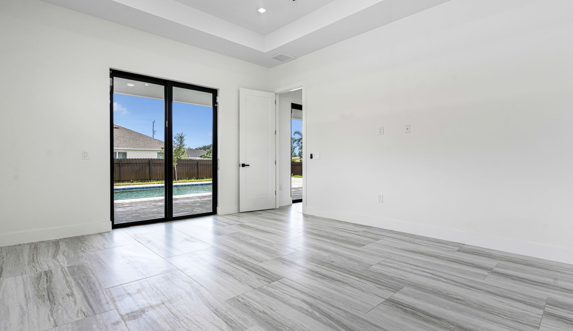 Empty white room with large windows overlooking a pool. Grey wood-like flooring and white door.