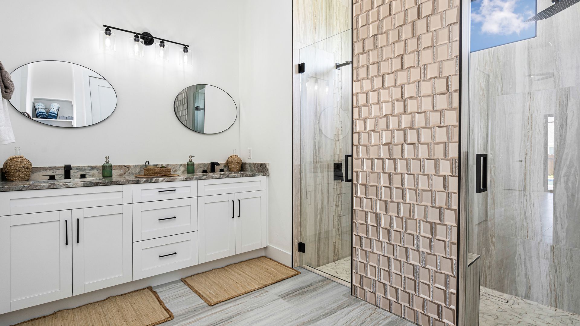 Bright bathroom with white cabinets, dual sinks, patterned shower wall, and oval mirrors.