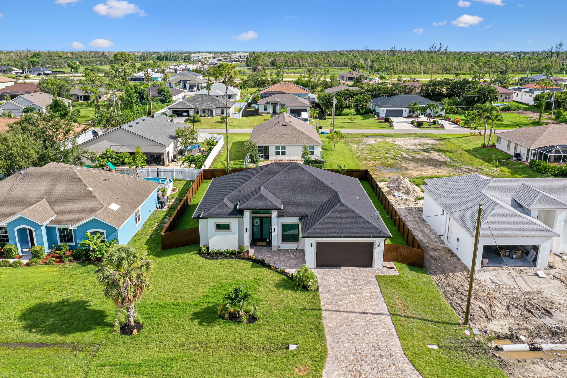 Aerial view of suburban homes, the closest with a dark roof, tan driveway, and green yard.