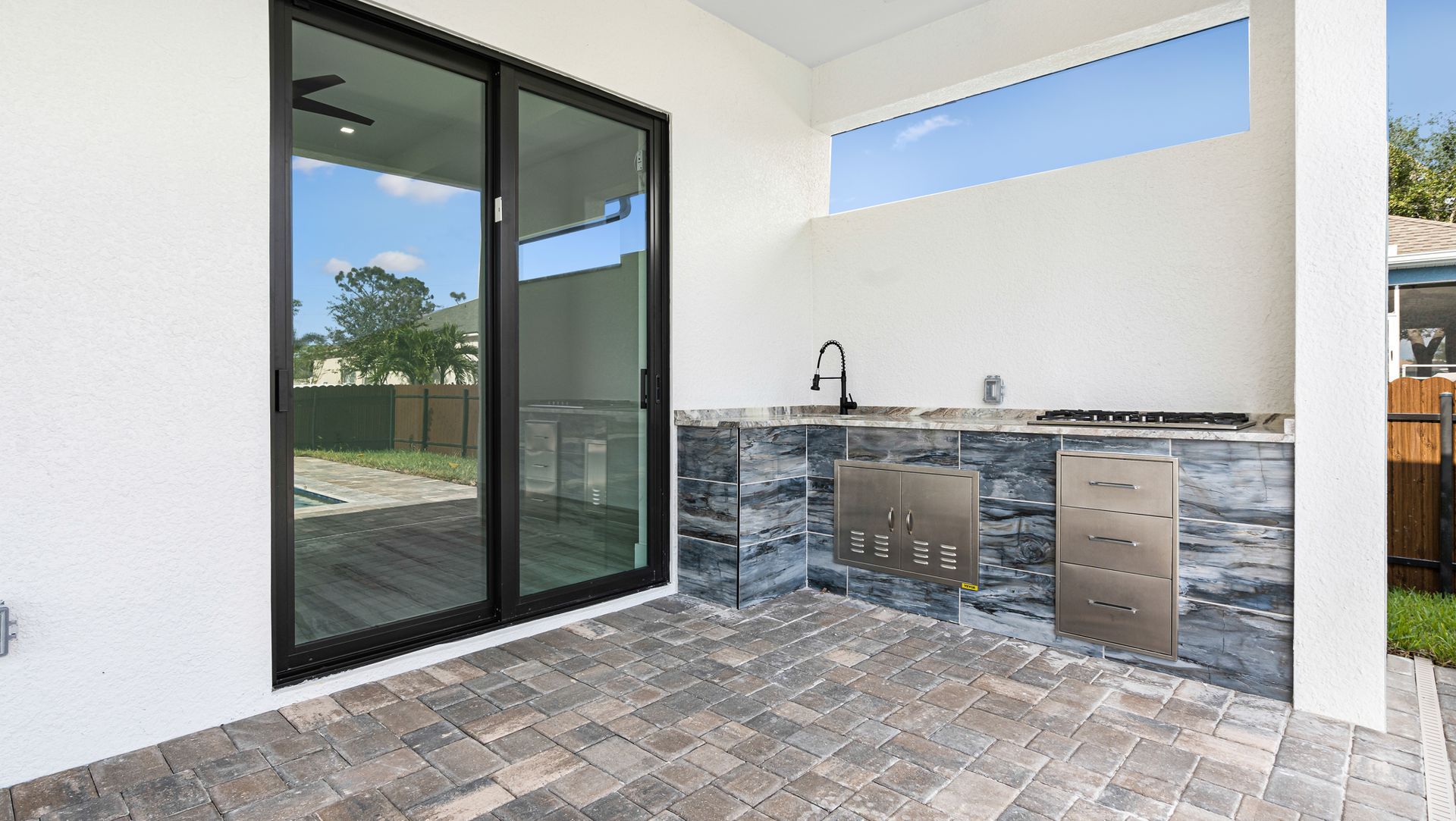 Outdoor kitchen with grill, sink, and sliding glass doors. Grey stone counter and flooring.