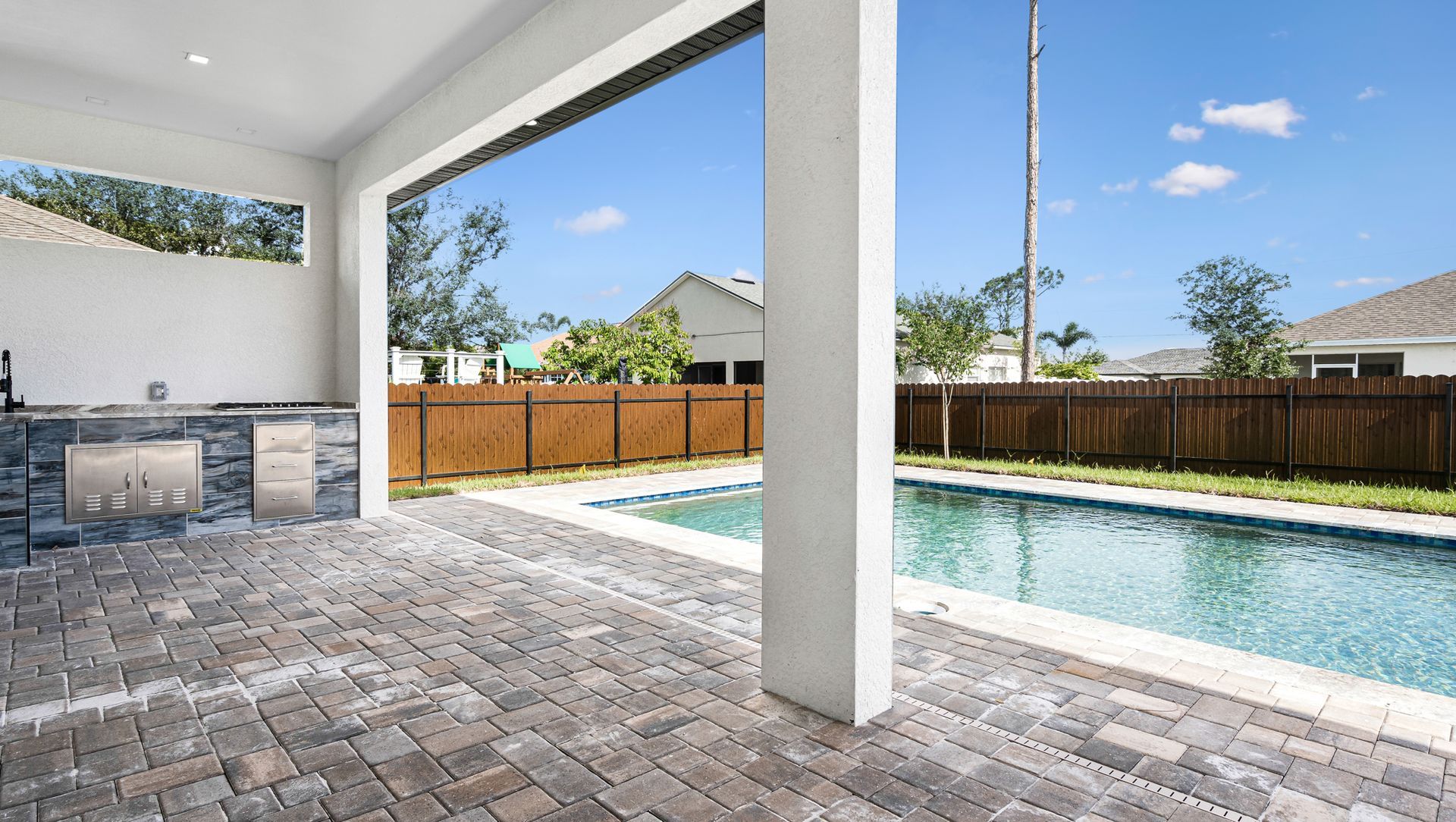 Patio with outdoor kitchen, pool, and blue sky. Brick flooring, white columns, brown fence.