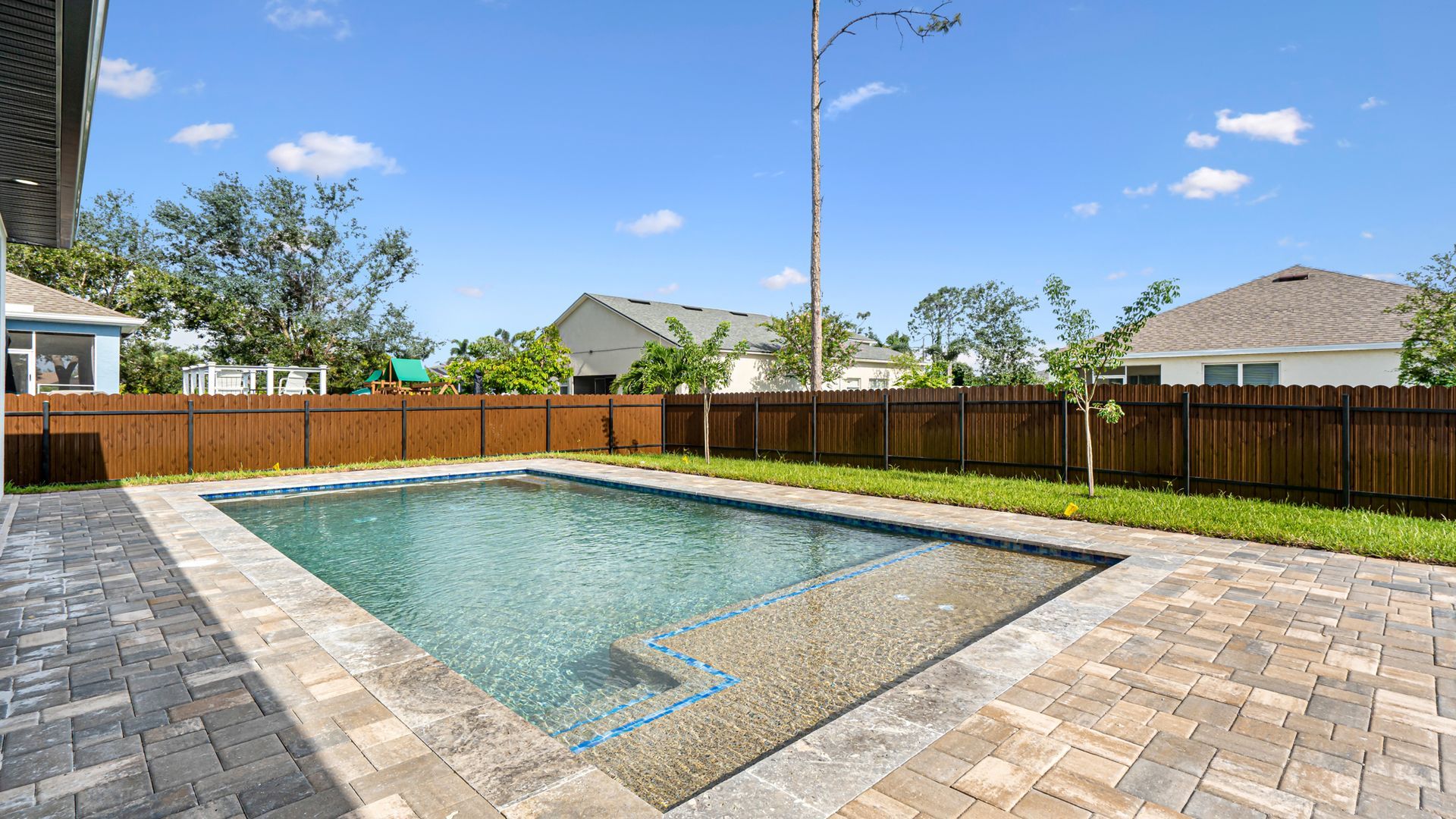 Backyard pool with brick patio, fenced yard, and blue sky.