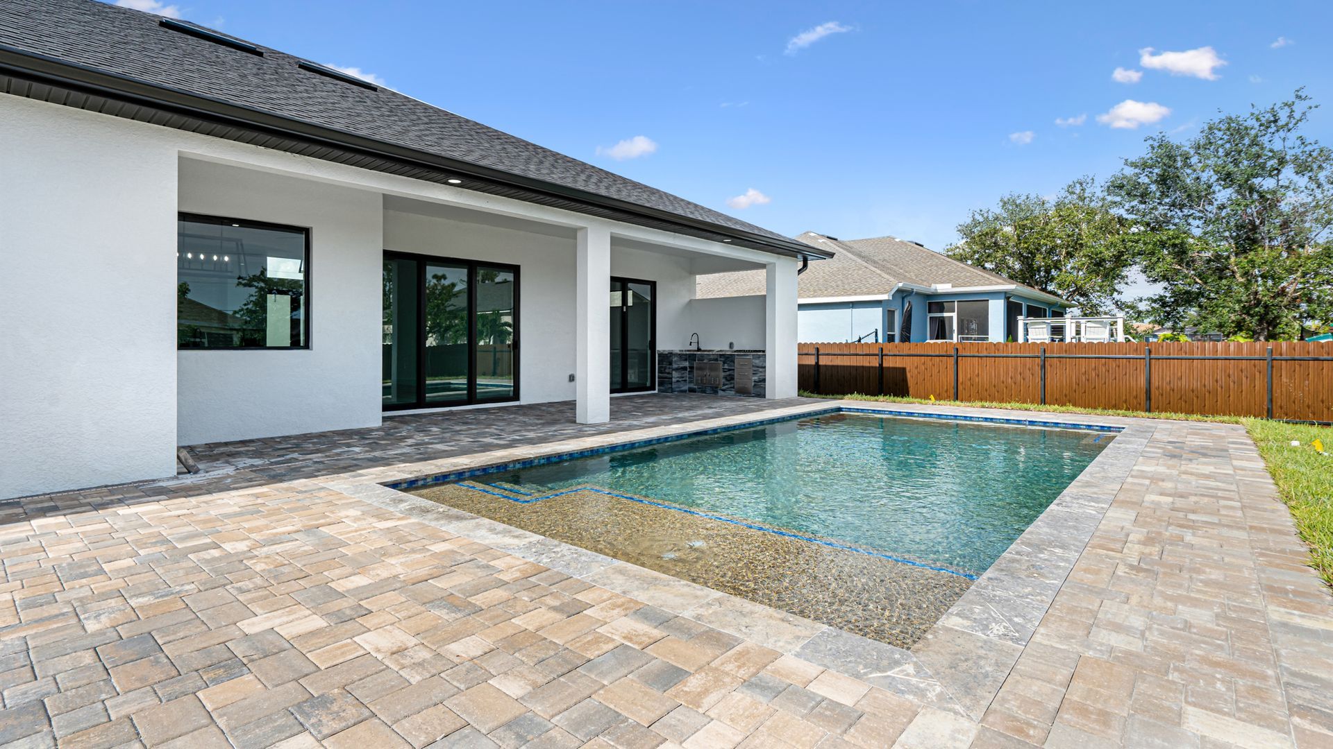 Backyard with pool and patio. White house with black trim, blue sky, and green lawn.