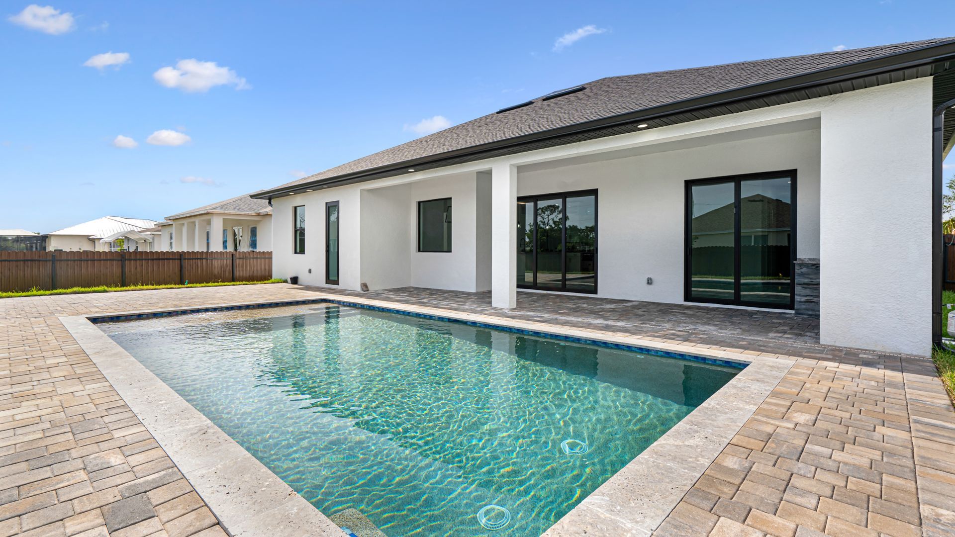 Backyard with pool, light stucco home, black window frames, and brick patio on a sunny day.
