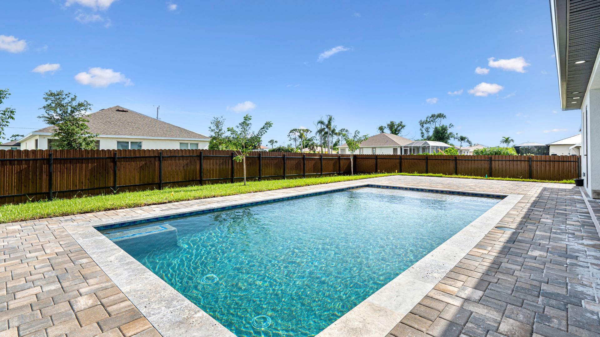 A rectangular swimming pool surrounded by a brick patio and a wooden fence under a blue sky.