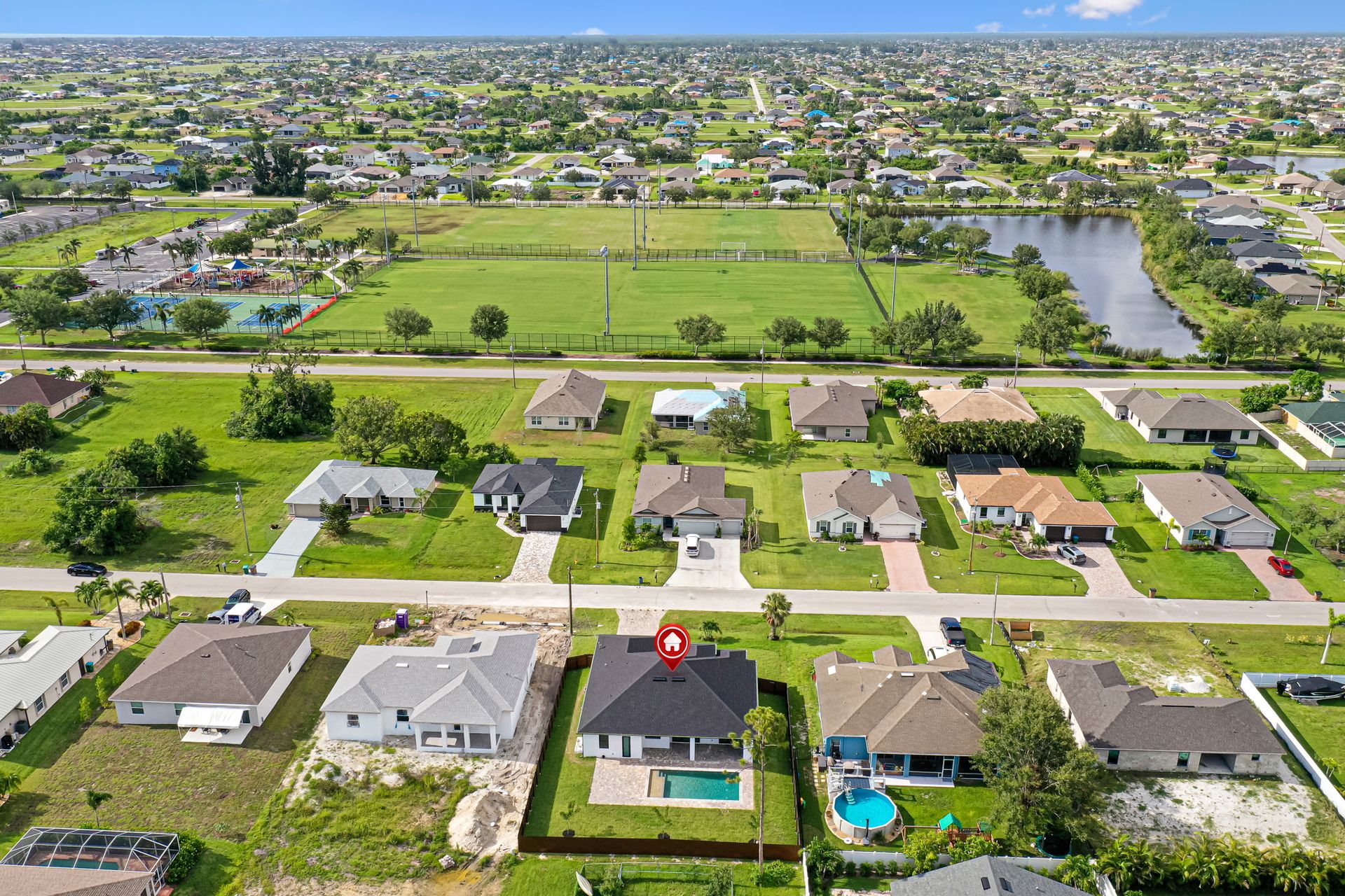 Aerial view of suburban houses with a central black-roofed house and a pool. Green field and lake in the background.