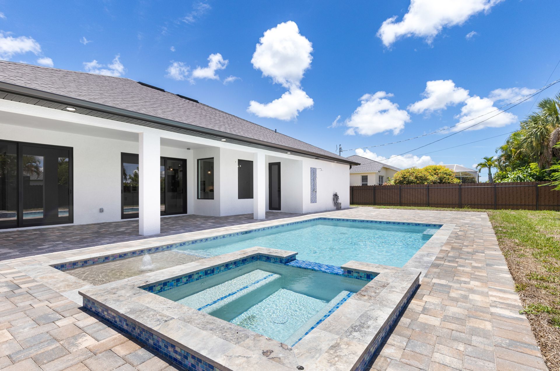 Backyard with a pool and spa, white house, blue sky, and green grass.
