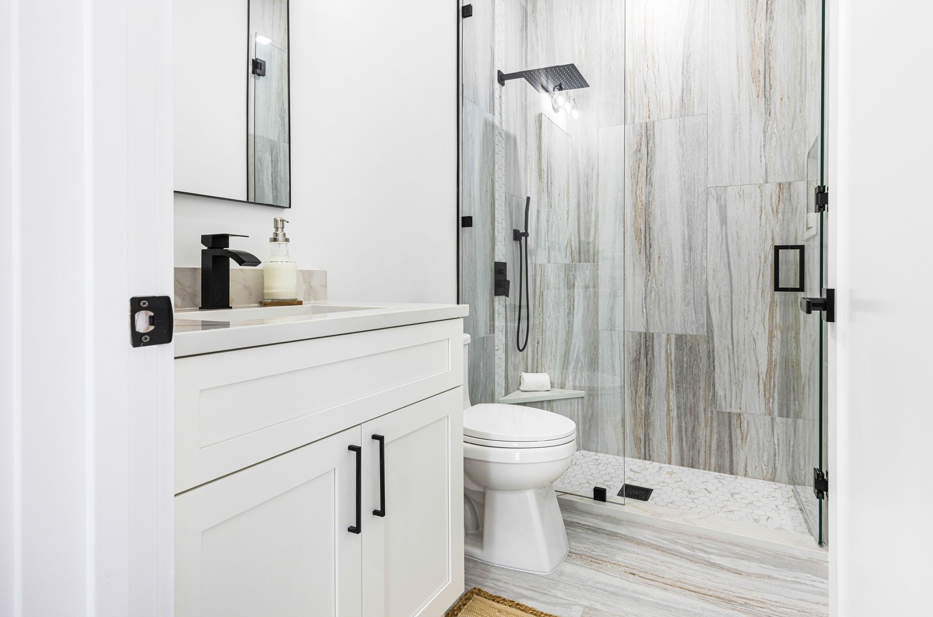White bathroom with a glass shower, vanity, and toilet. Black fixtures and light-colored stone tiles.