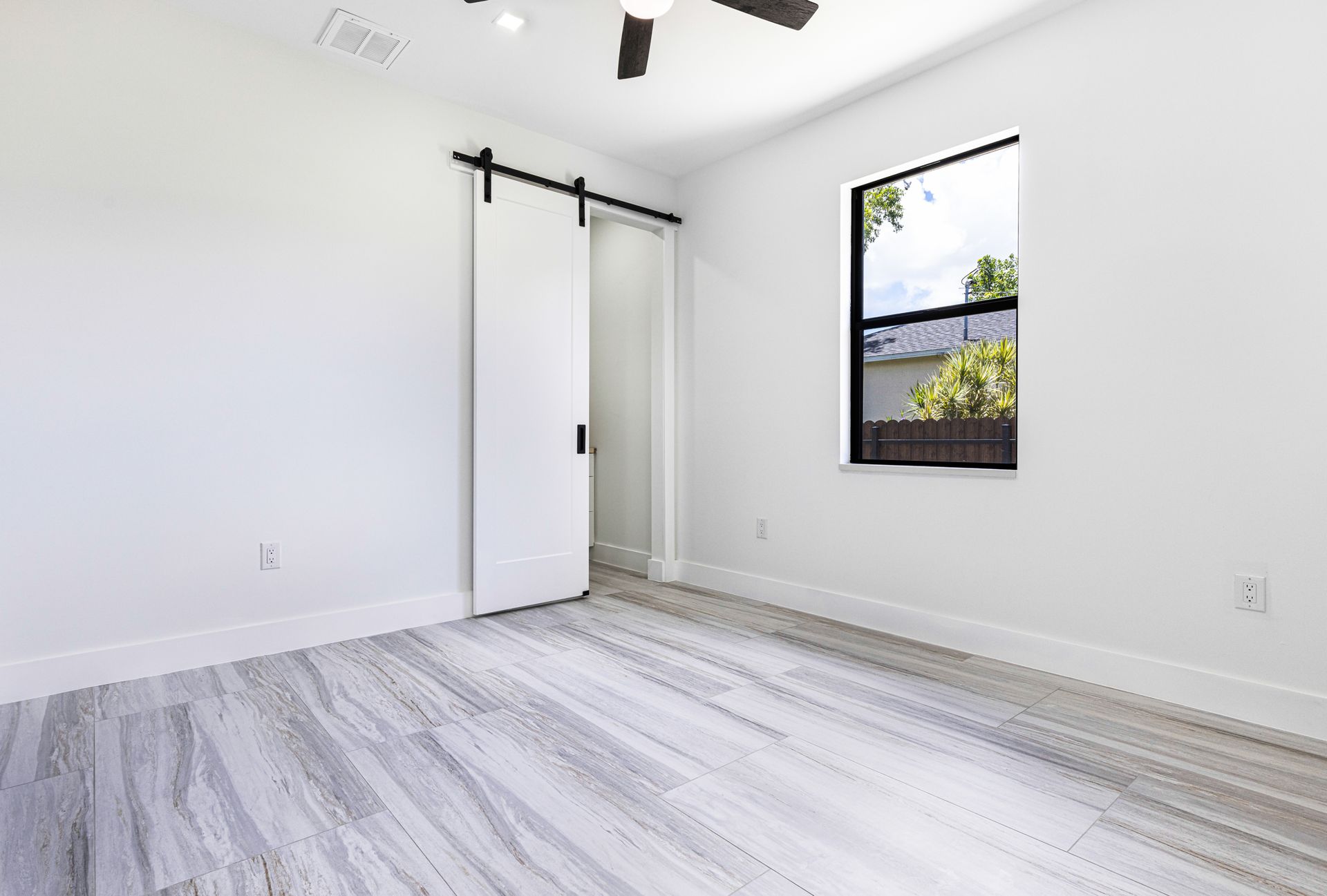 Empty white bedroom with light wood-look flooring, barn door, and a window with an outside view.