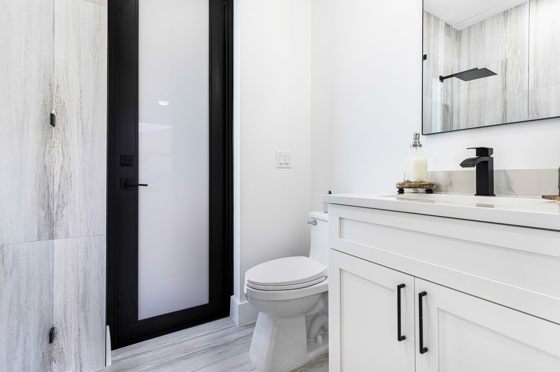 Modern white bathroom with black door, toilet, vanity, and shower.