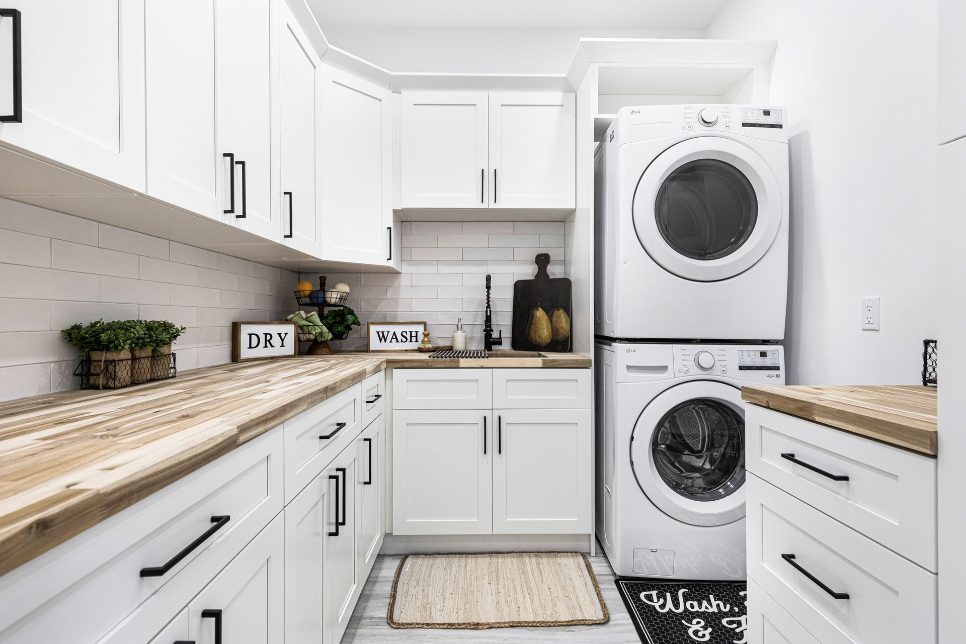 White laundry room with stacked washer/dryer, wooden countertops, white cabinets, and black hardware.