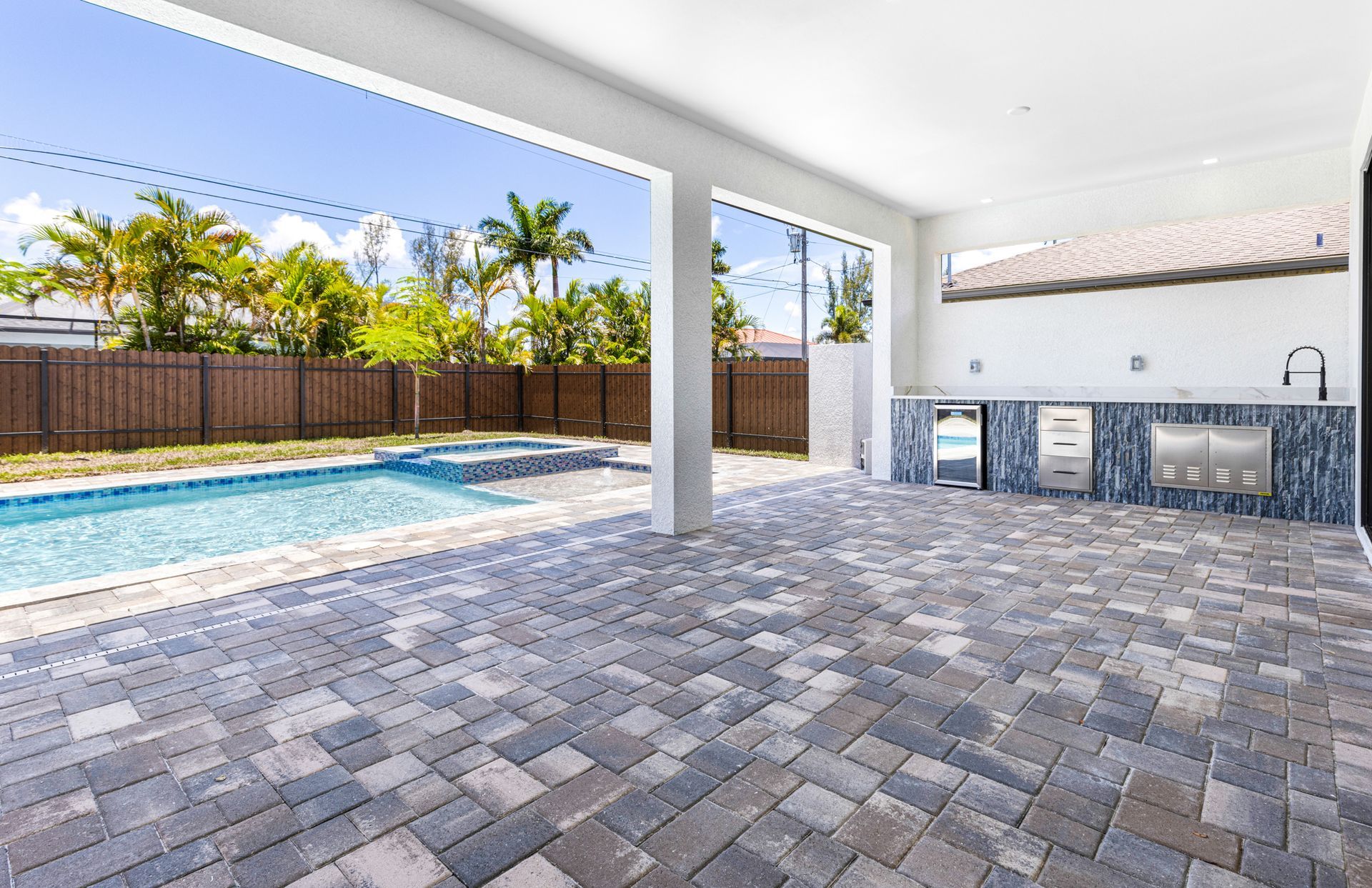 Patio with pool, outdoor kitchen, and paved floor, blue sky, palm trees and a wooden fence.