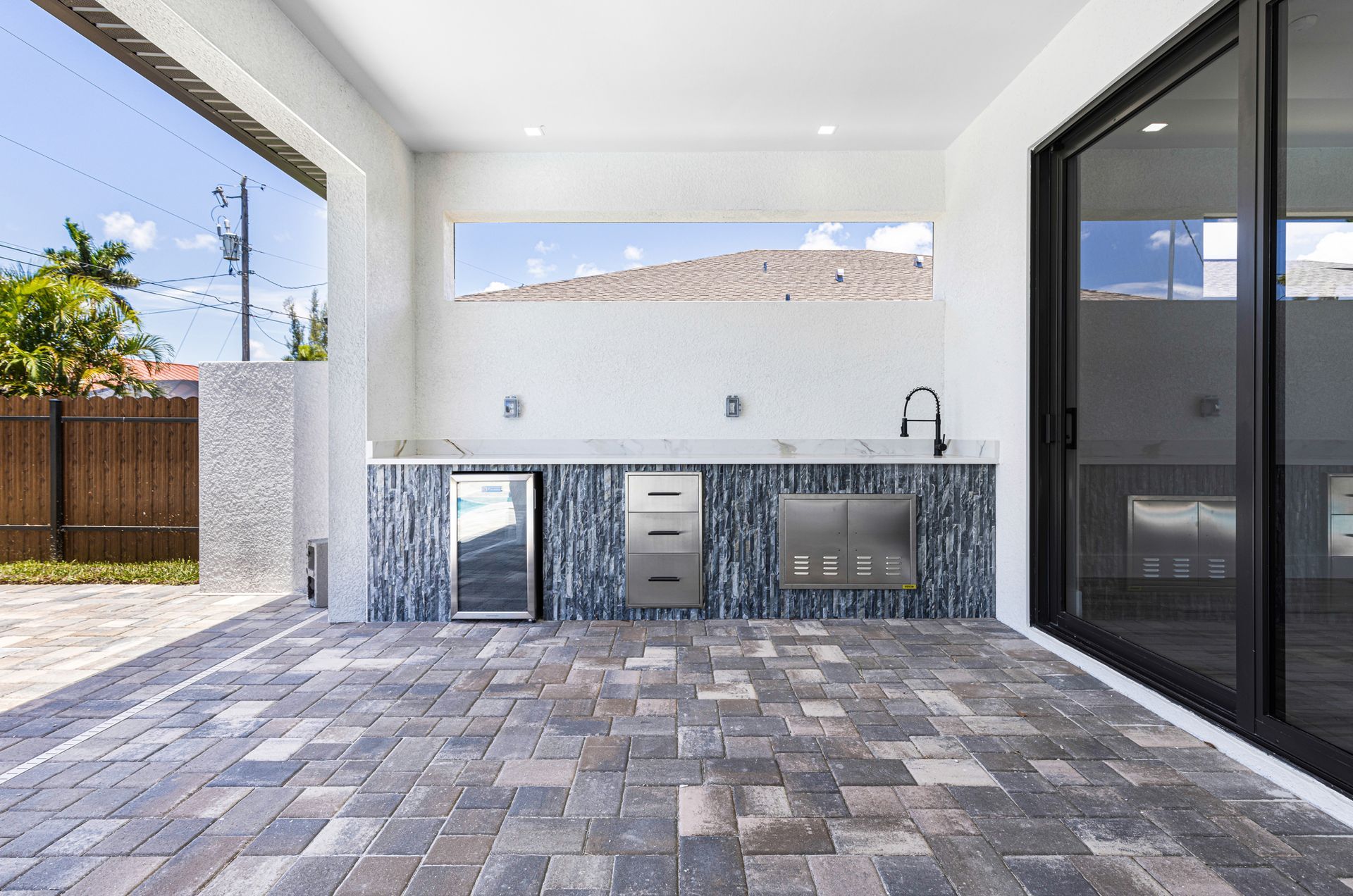 Outdoor kitchen with tiled backsplash, grill, sink, fridge, and brick patio.