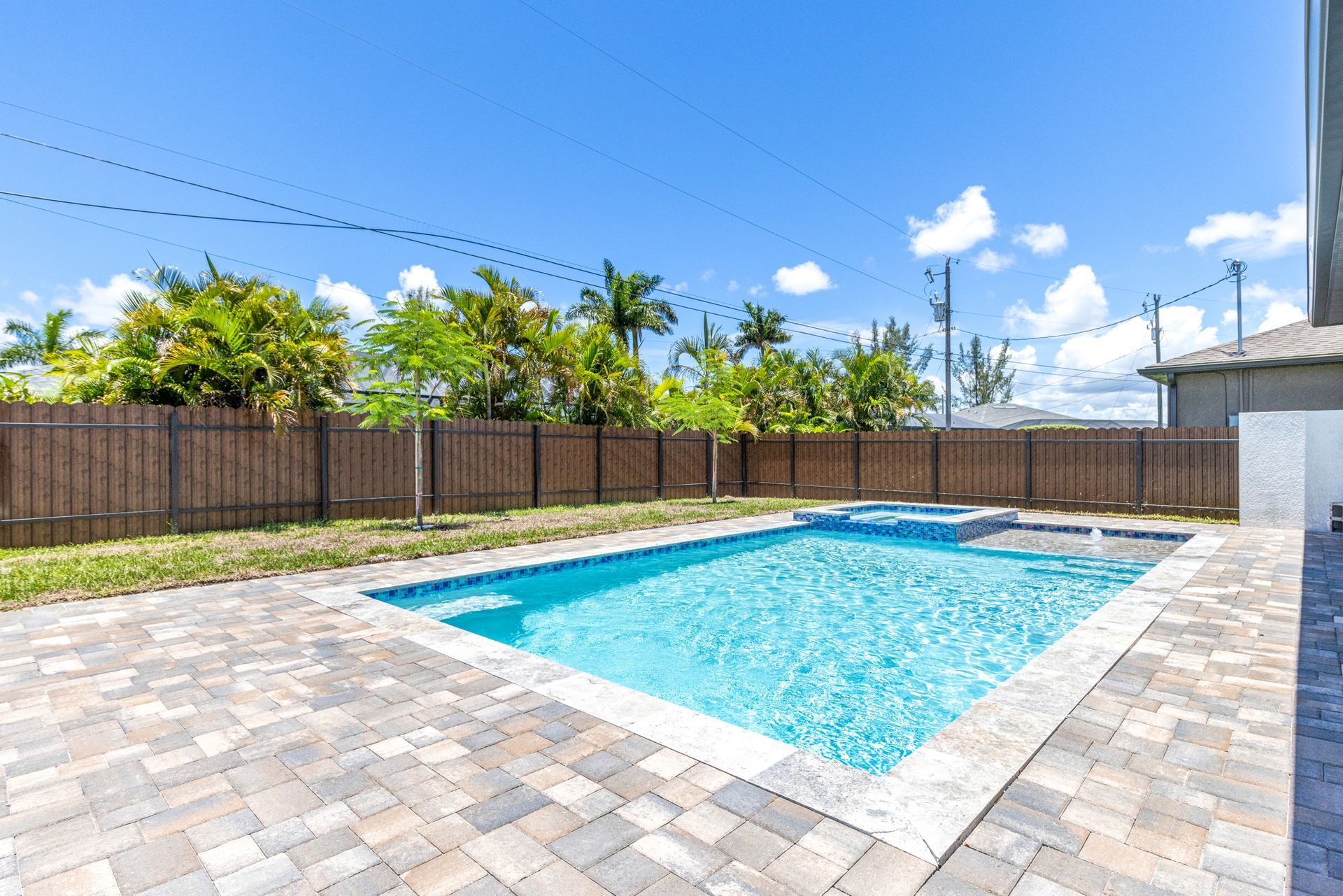 Backyard with a blue pool, hot tub, brick patio, and wooden fence under a blue sky.
