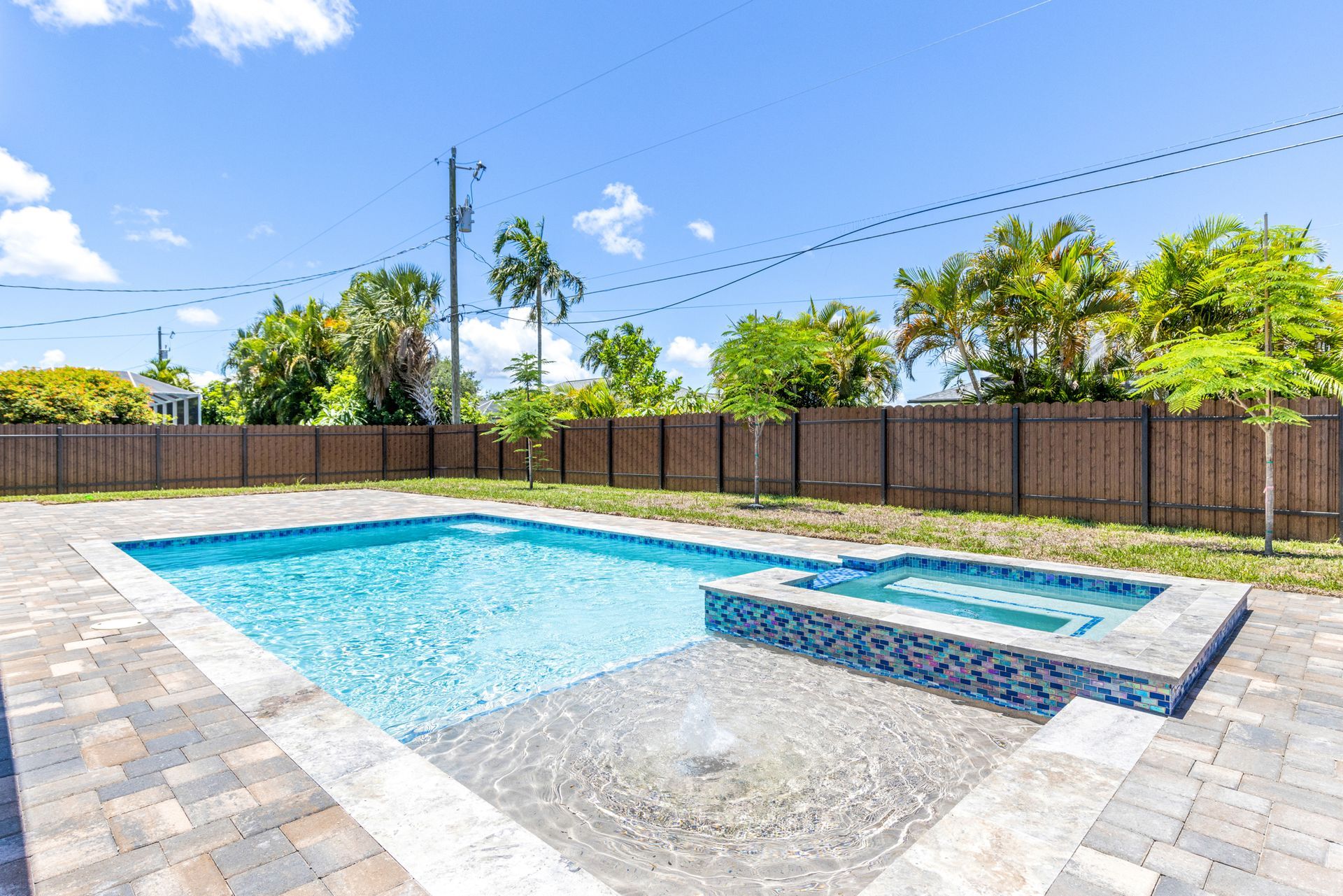 Backyard with pool and hot tub, surrounded by a brick patio and wooden fence, under a sunny, blue sky.
