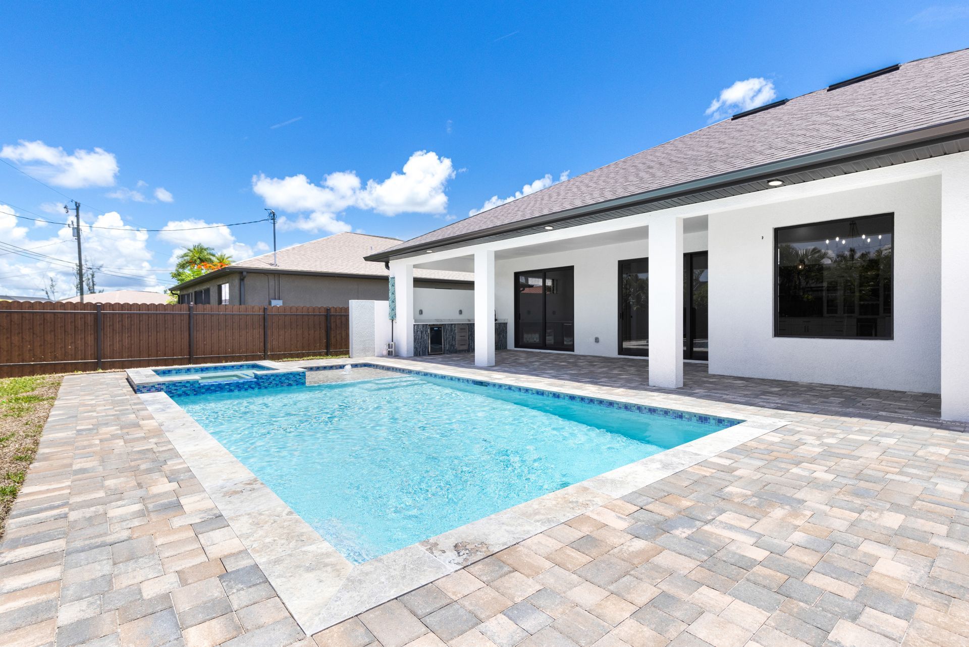 Backyard with pool and jacuzzi, white house, blue sky, and paver patio.