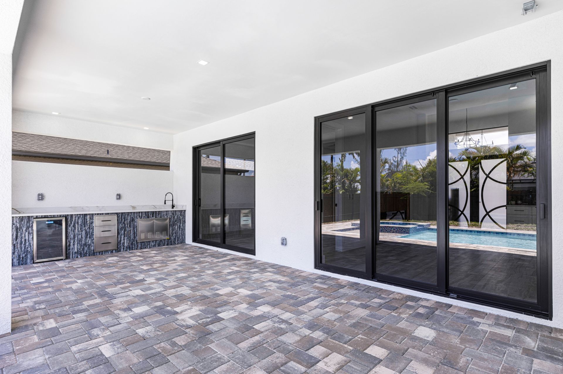 Outdoor patio with a kitchen, sliding glass doors, and a brick floor. Pool visible through the doors.