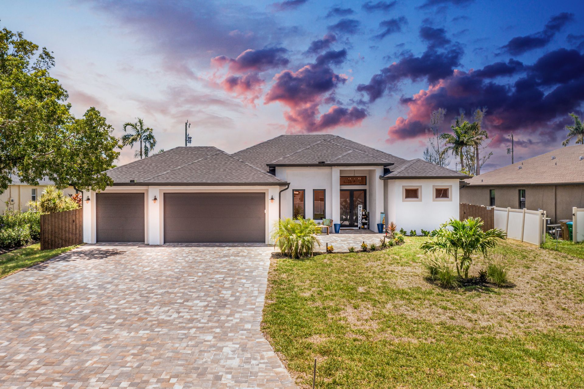 Modern white house with brown garage doors, driveway, and tile roof under a colorful sky.