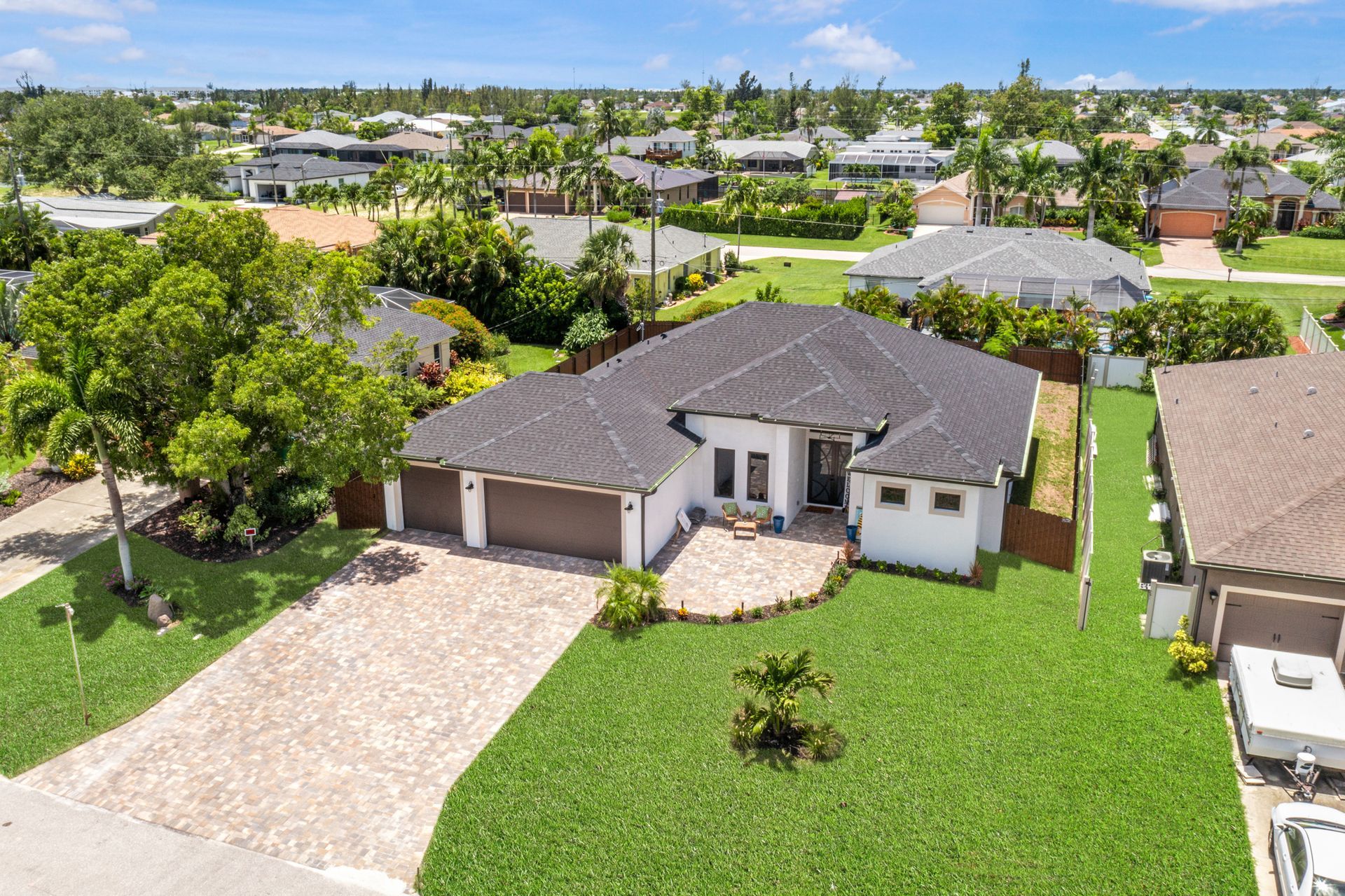 Aerial view of a white house with a dark roof, brown garage doors, and a stone driveway, surrounded by green grass and trees.