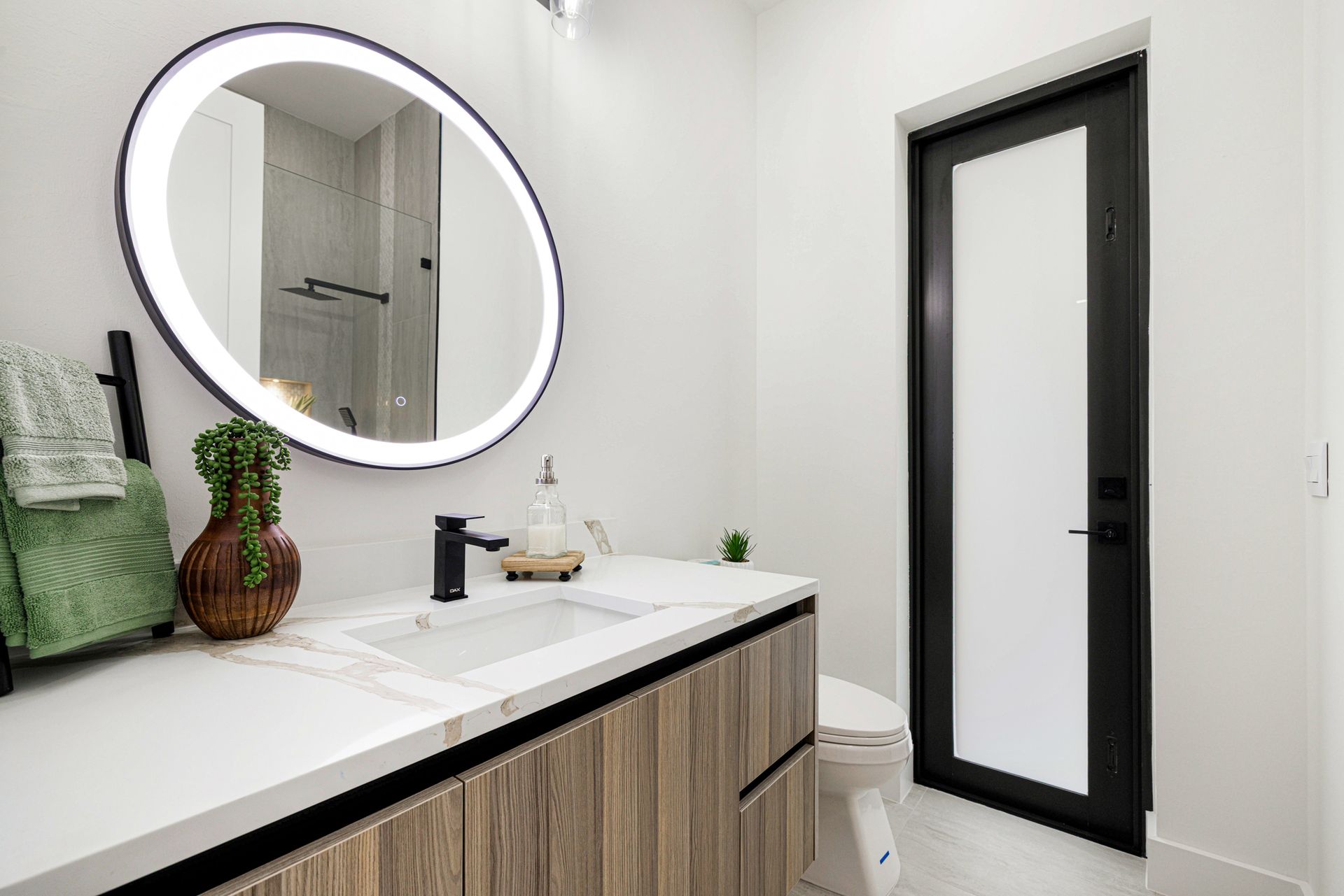 Bathroom with a round, illuminated mirror, black faucet, wooden vanity, and door.