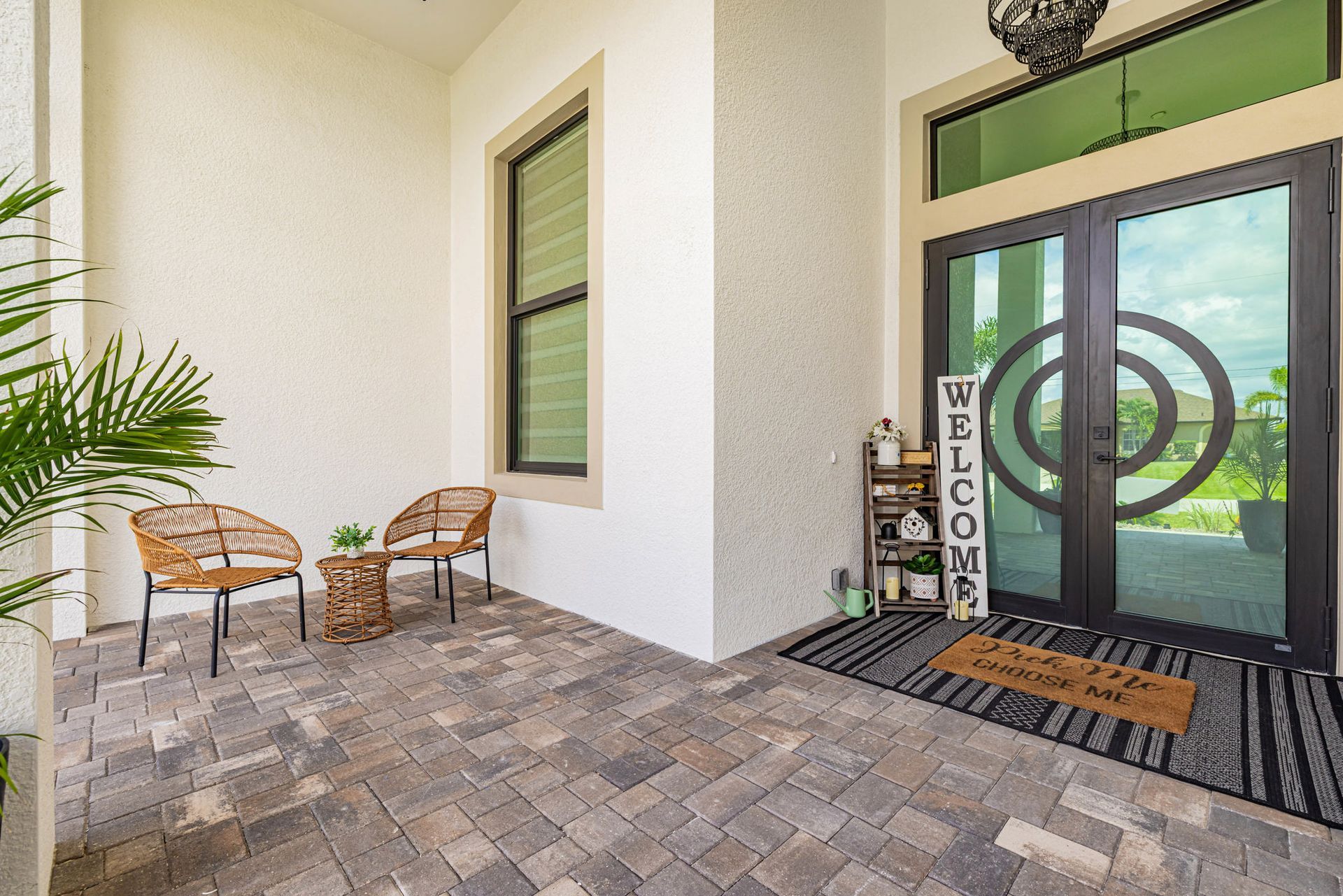 Entryway with seating, a welcome sign, and double glass doors. Paved patio in front of a white stucco wall.