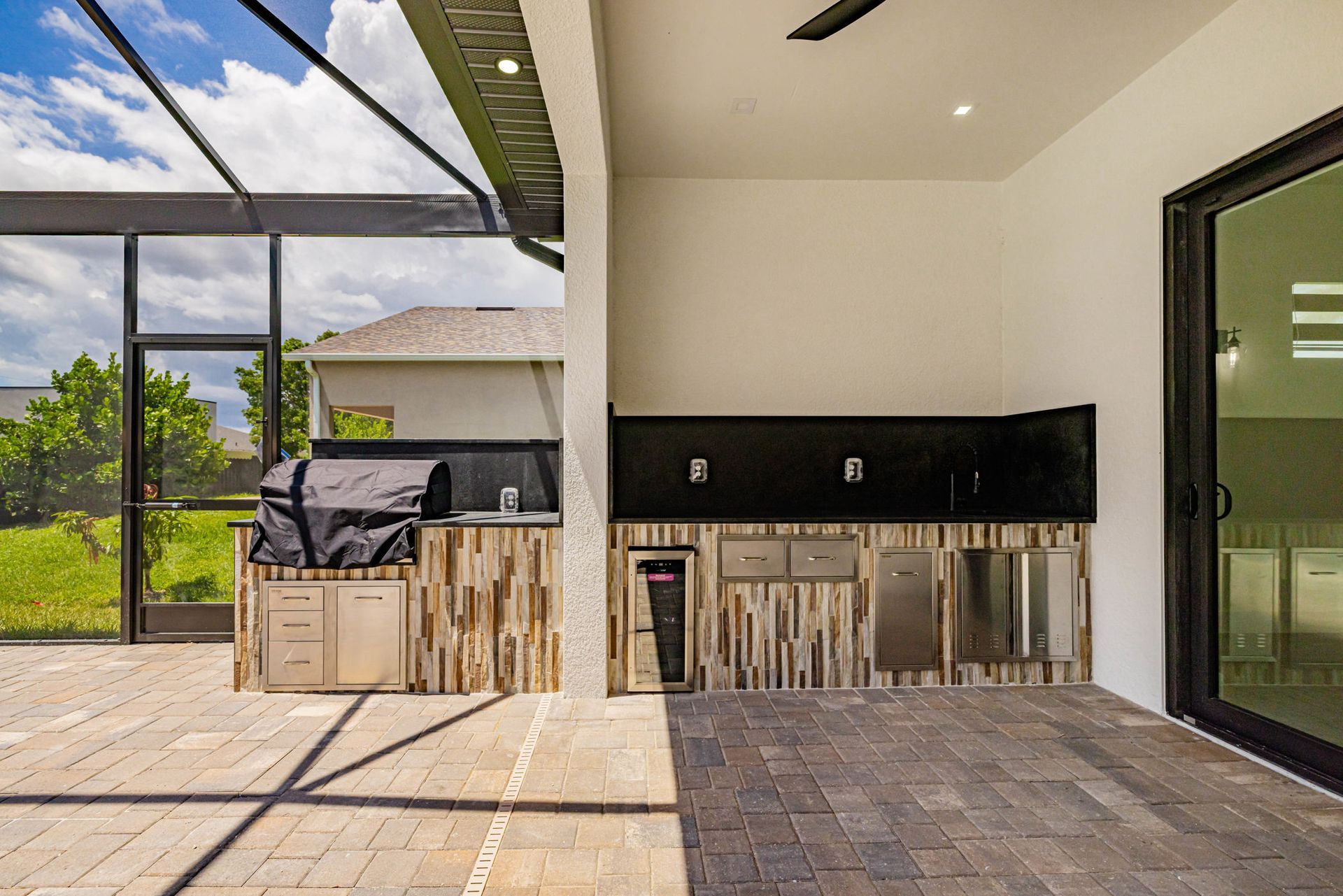 Outdoor kitchen with grill, cabinets, and a wine cooler on a brick patio under a screened enclosure.